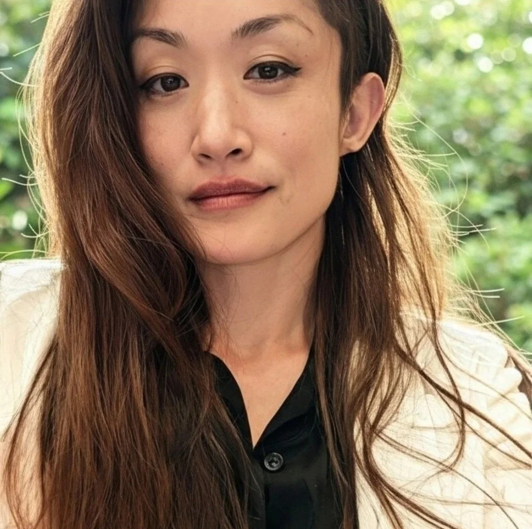 Close-up of a woman with long, wavy brown hair, lightly flushed cheeks, light makeup, and wearing a black shirt with a white blazer, outdoors with green foliage in the background.