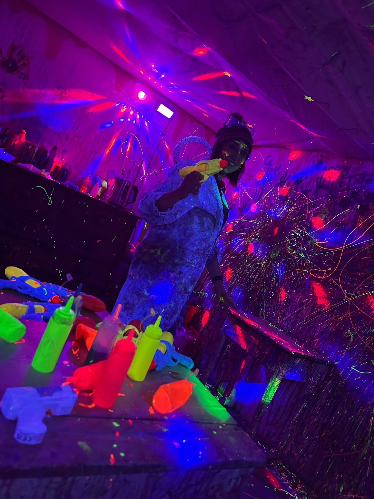 Person holding a yellow toy with a red nose, in a room with glowing paint, fluorescent bottles, and colorful light effects.