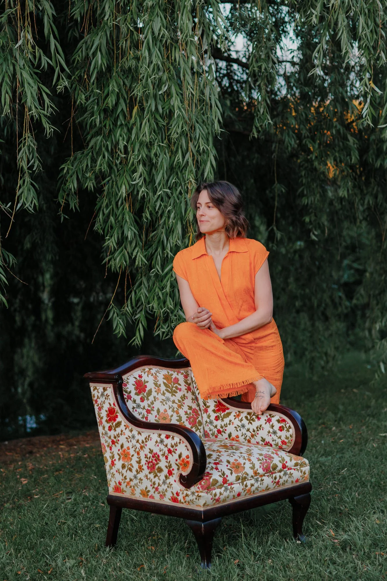 Montreal podcast host in an orange jumpsuit, kneeling on a vintage floral armchair under a willow tree