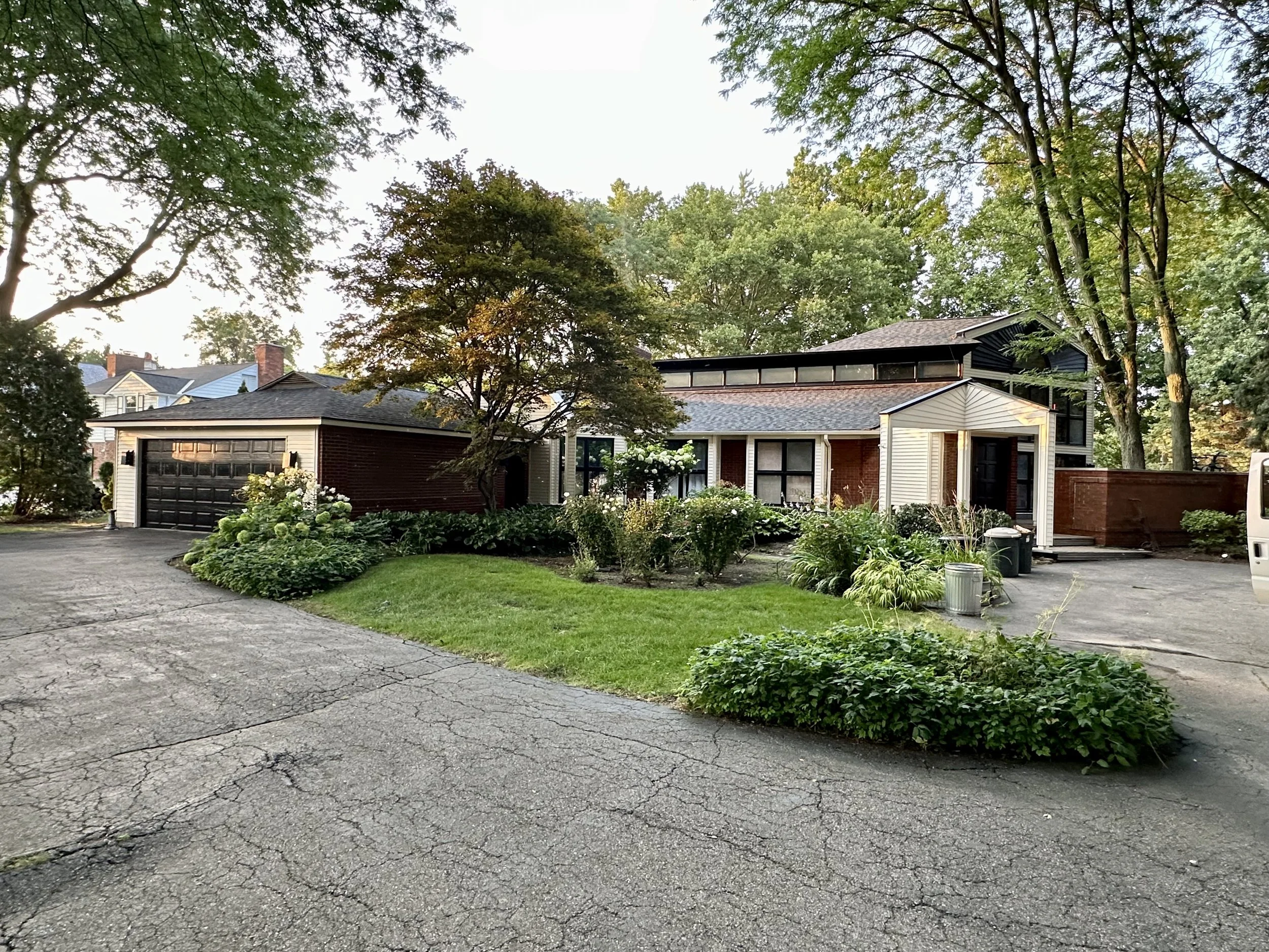 A residential house with a brick and siding exterior, surrounded by trees and shrubs, with a driveway and a garage on the left.