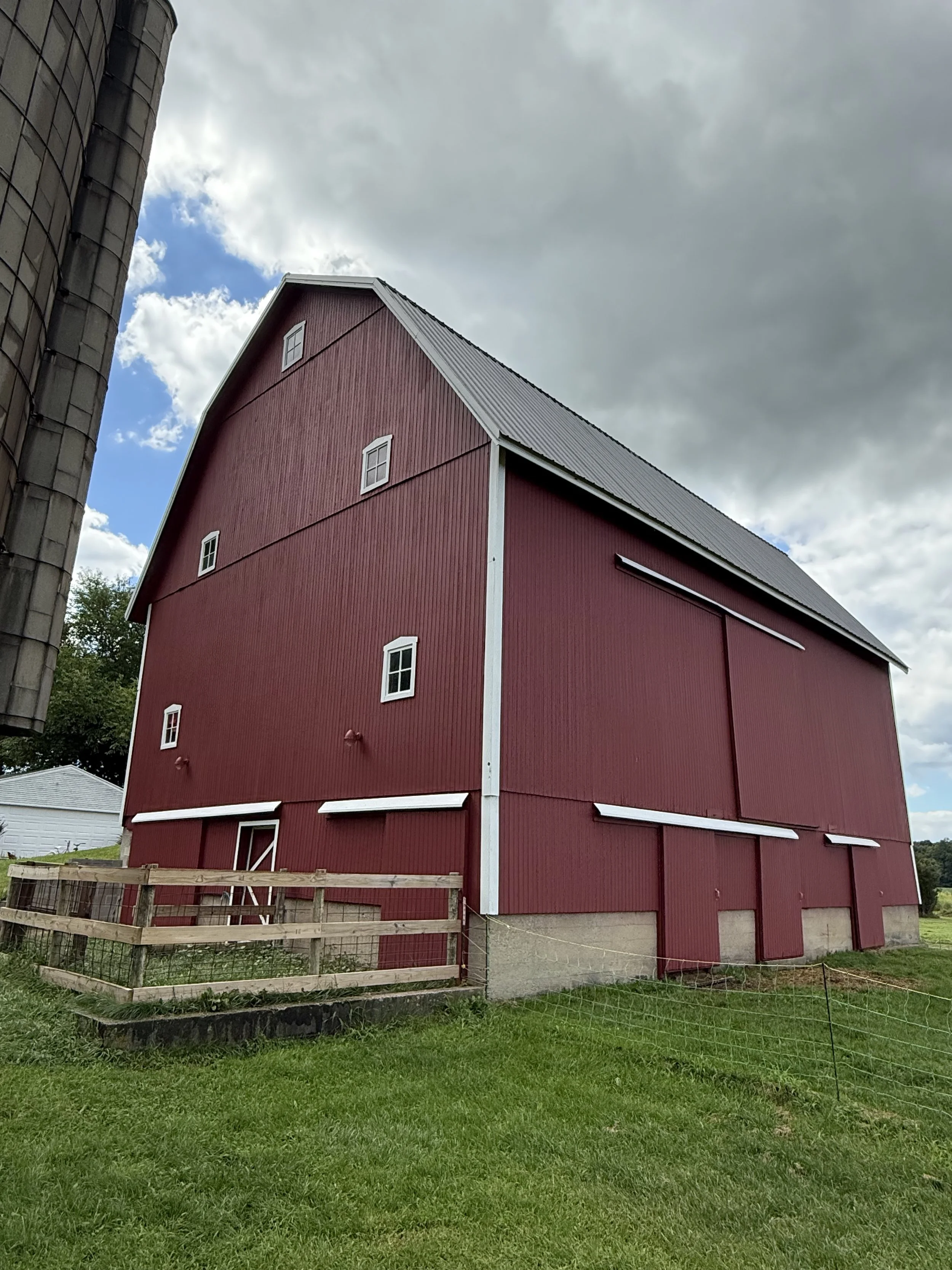 Red barn with small windows and a metal roof, surrounded by green grass and cloudy sky.