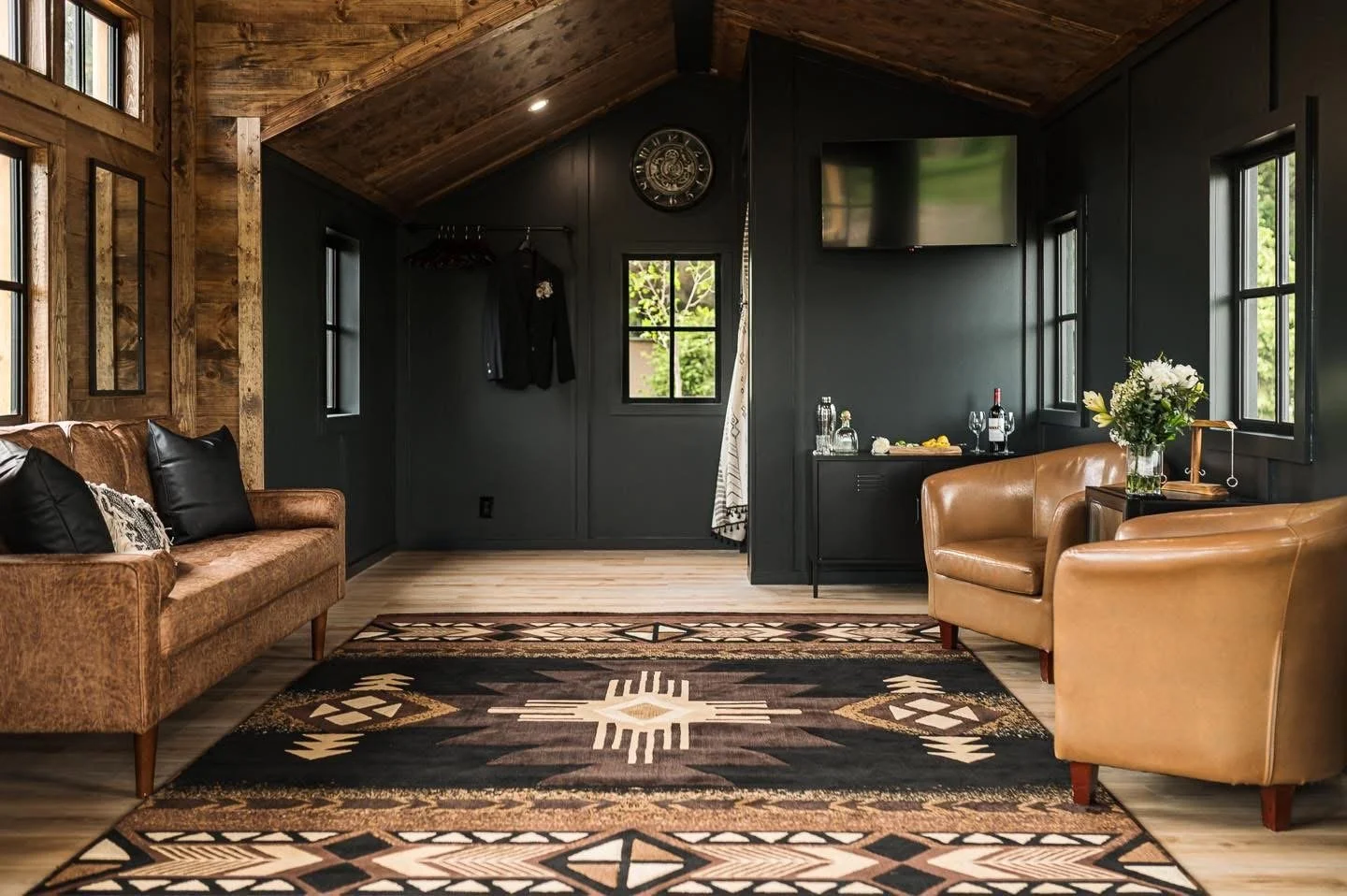 A living room with black walls, wooden ceiling, and hardwood floor. Features a brown leather sofa with black pillows, two tan leather armchairs, a colorful patterned rug, a wall-mounted TV, a small black sideboard with water bottles, glasses, and a vase with white flowers, and windows letting in natural light.