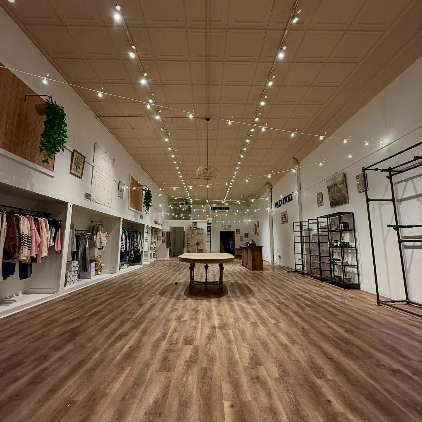 An empty clothing boutique with wooden flooring, racks of clothes along the left wall, black metal shelves on the right, and string lights hanging from the ceiling.