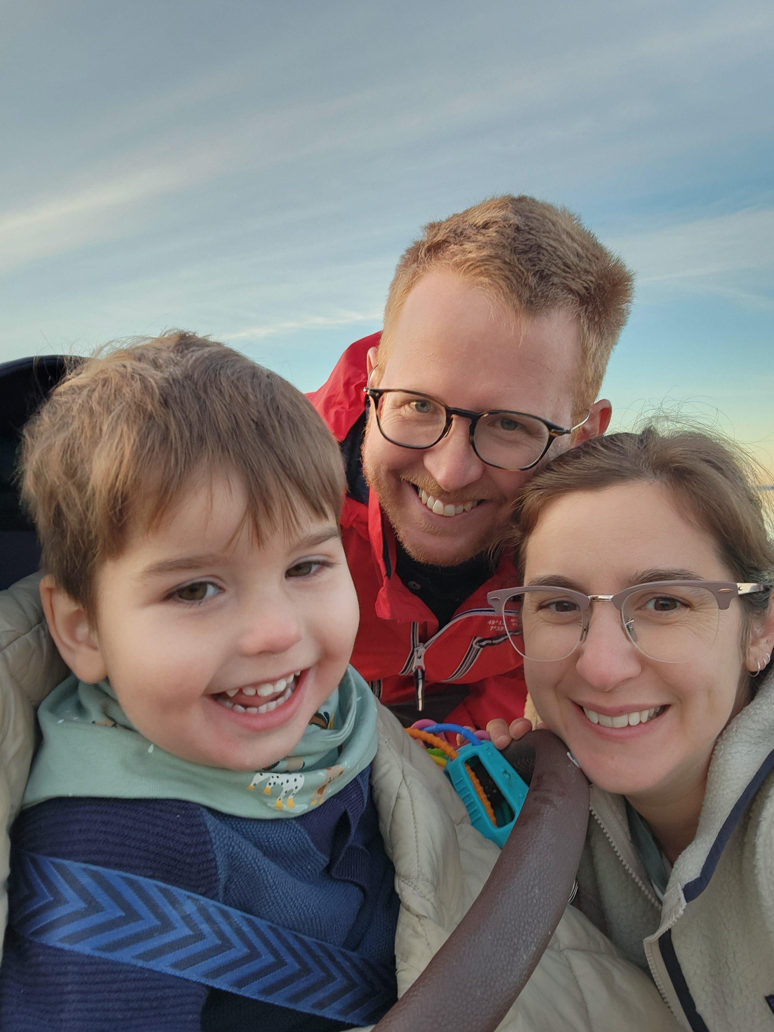 A smiling family selfie outdoors with a young child bundled in a stroller, two adults, one wearing glasses and a red jacket, taking the photo under a clear sky.