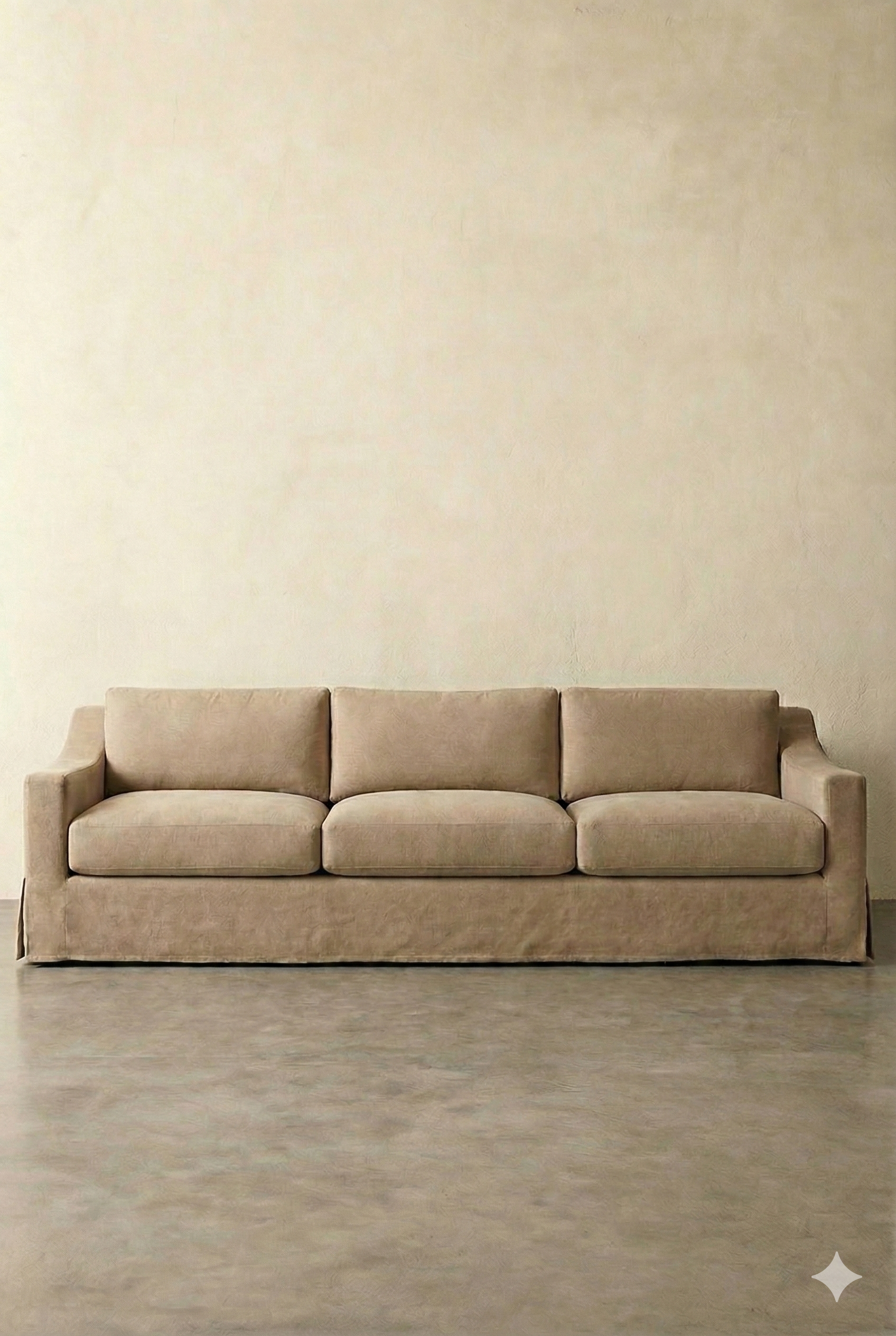 A beige sofa with four cushions against a plain wall on a concrete floor.