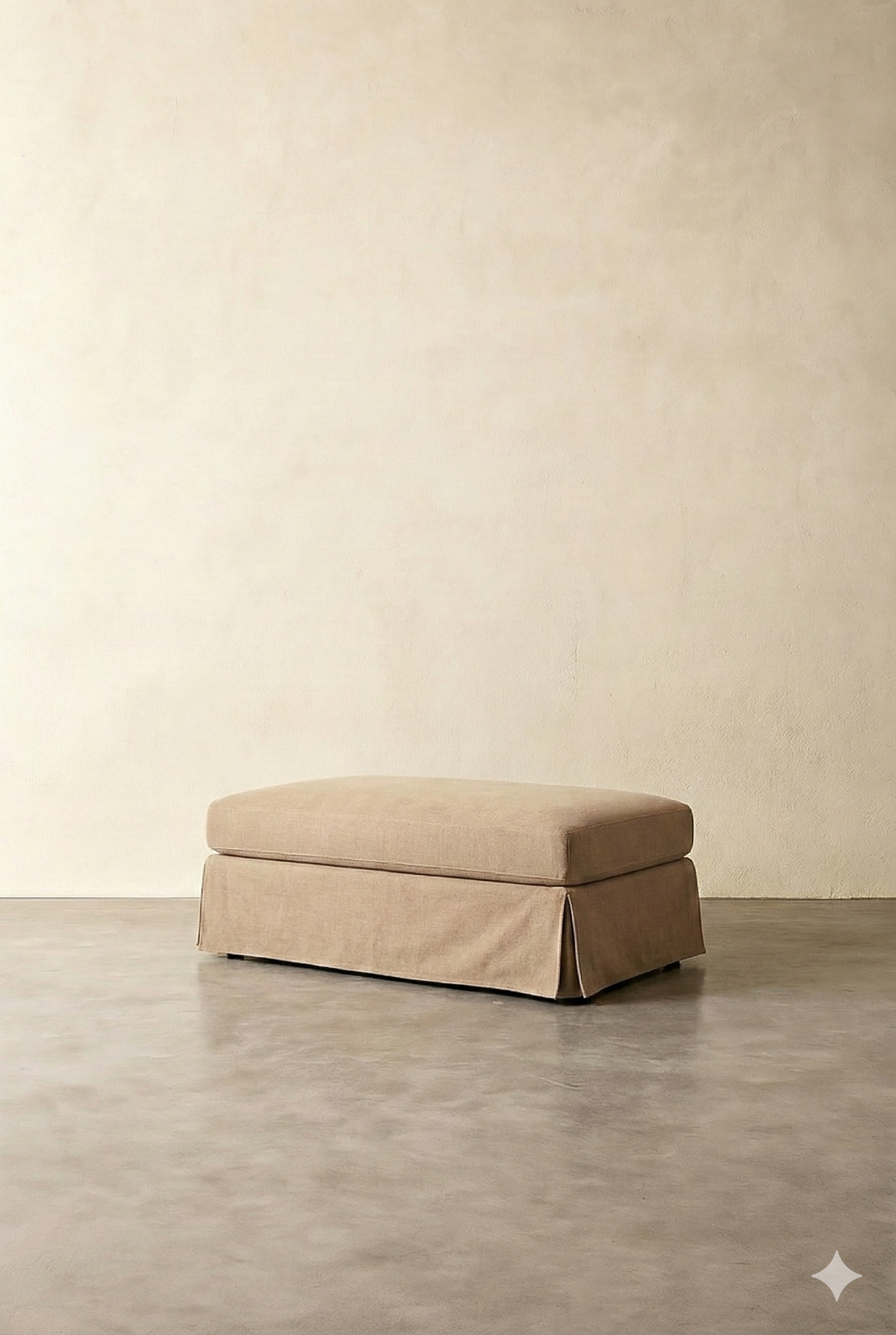 A beige upholstered ottoman with a skirted base, placed on a gray floor against a plain light-colored wall.