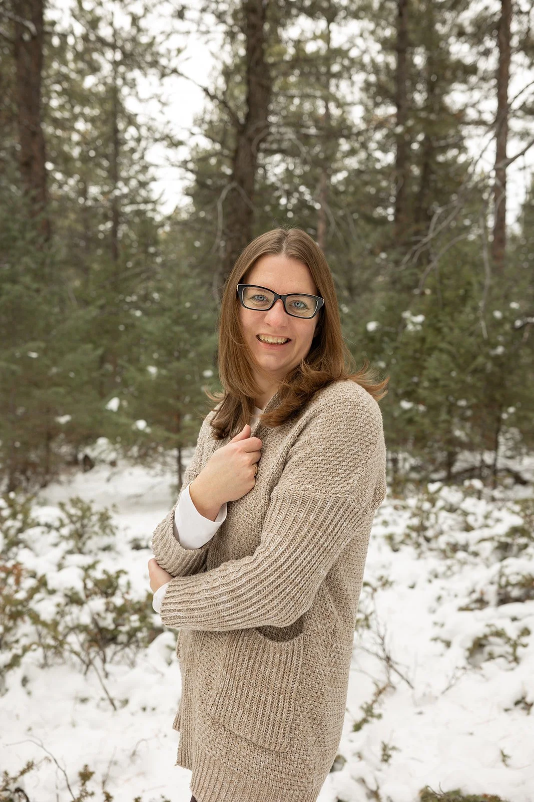 A woman with long brown hair, glasses, wearing a beige sweater and white shirt, standing outdoors in a snowy forest, smiling at the camera.