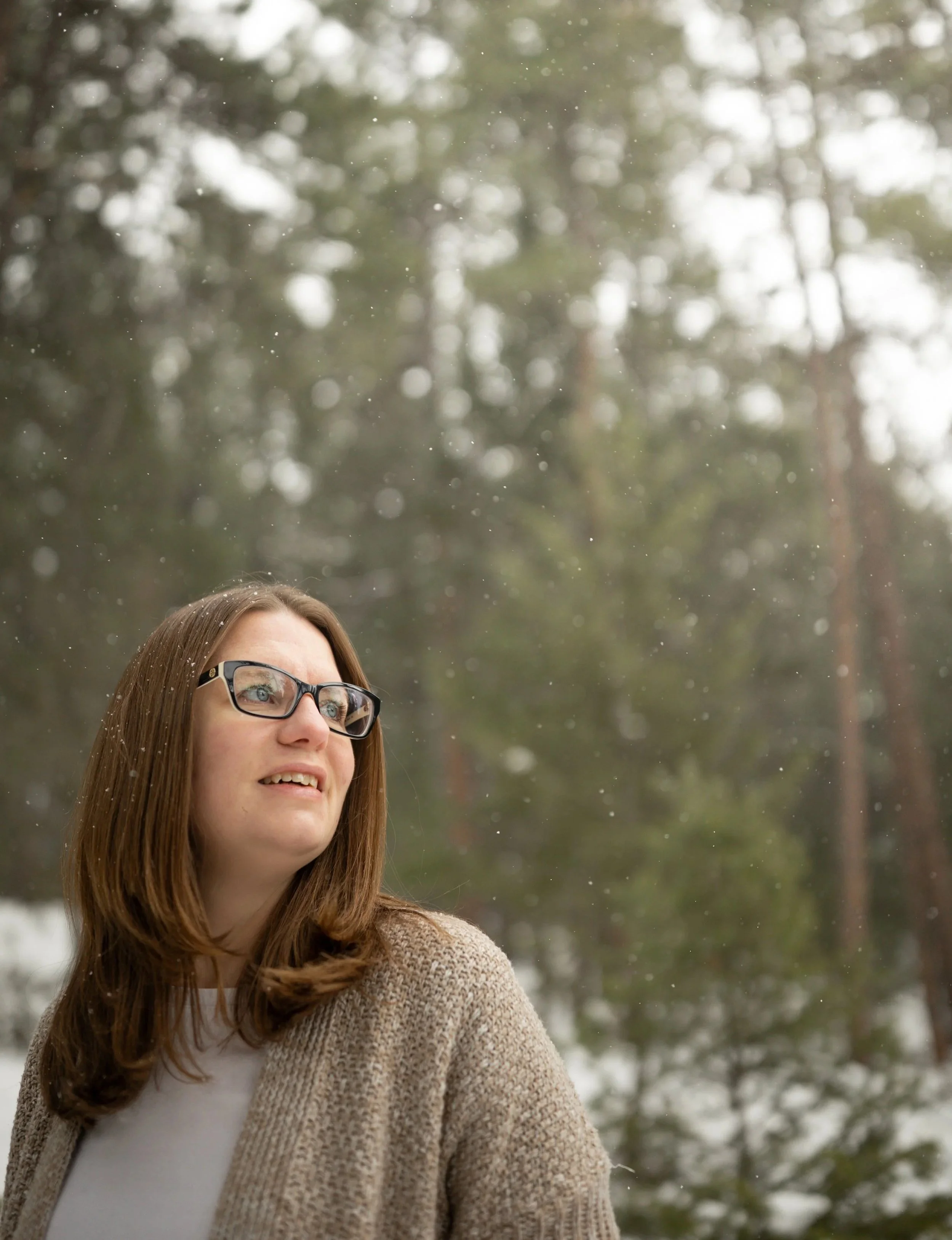 A woman with glasses and long brown hair looking up outdoors in a wooded area with snow and trees. Photo by Blue Flame Studio Co.