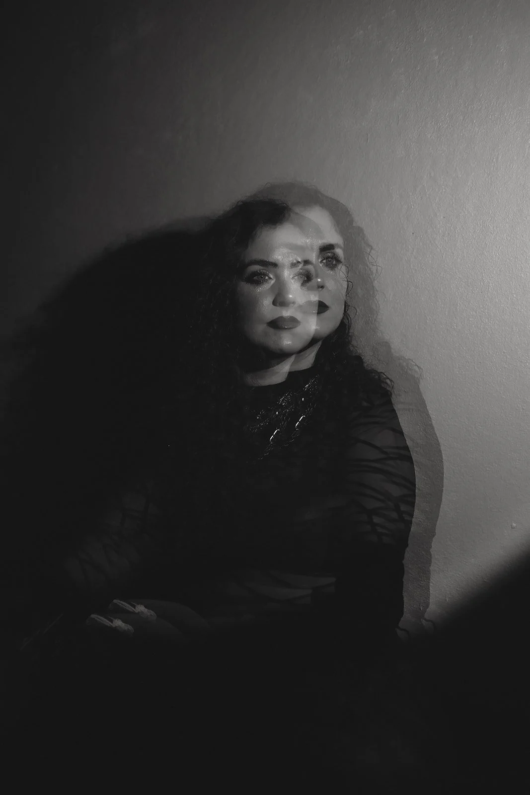 Black and white photo of a woman with curly hair, multiple exposures creating a ghosting effect, sitting against a plain wall.