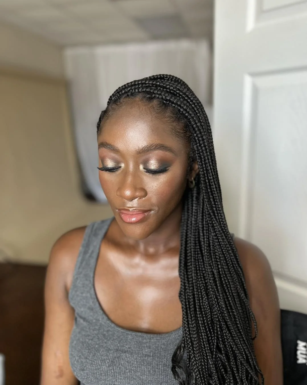A woman with braided hair and makeup, wearing a gray tank top, with her eyes closed and head tilted slightly down.