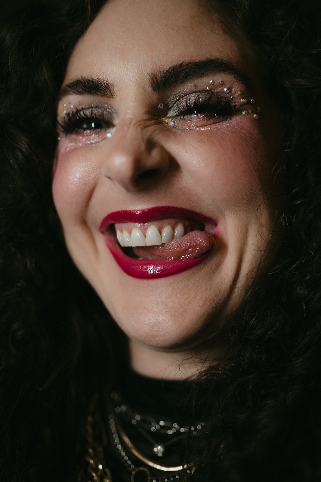 Close-up of a woman smiling with makeup, glitter, and rhinestones around her eyes, red lipstick, and her tongue sticking out.