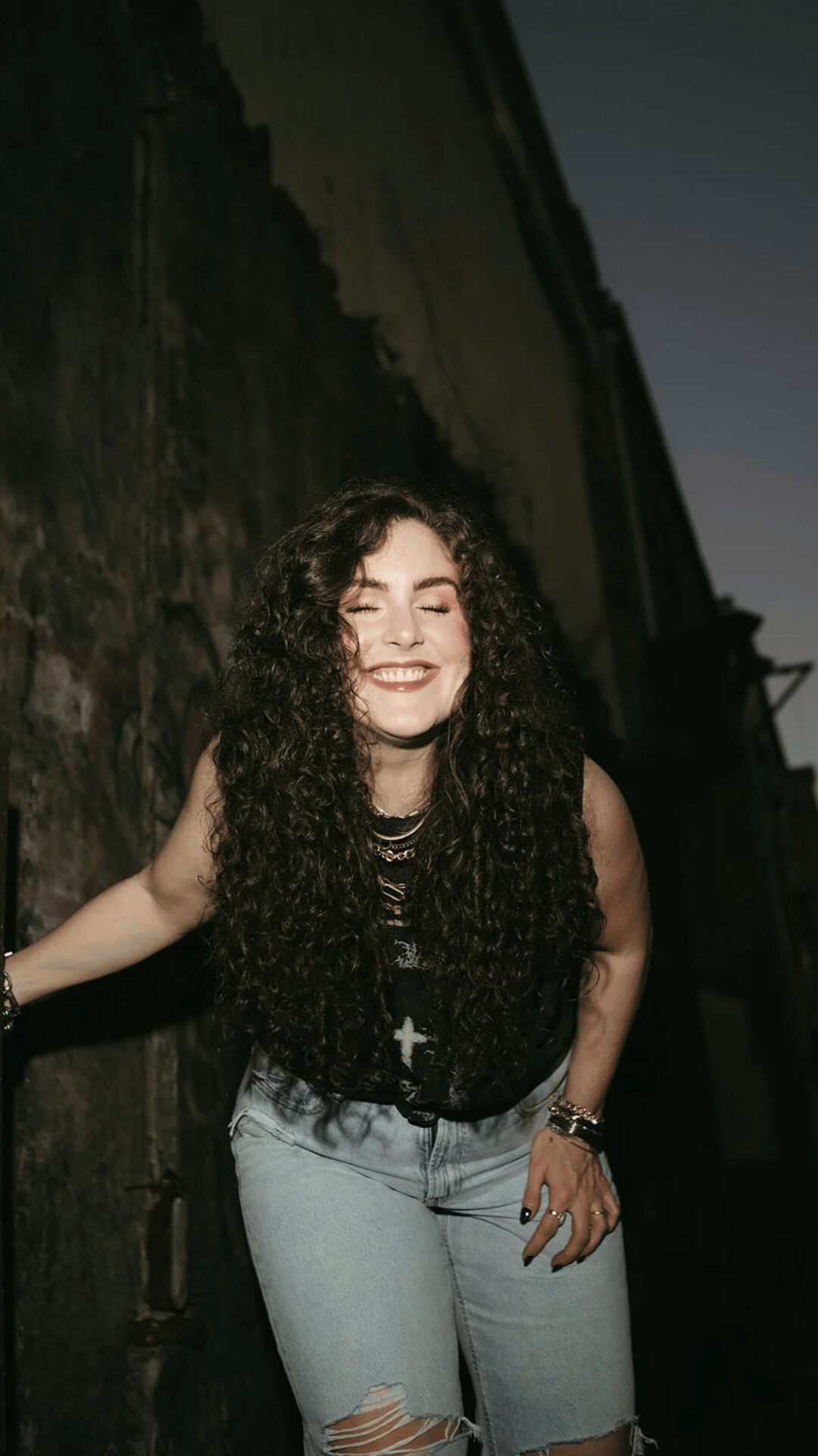 A young woman with long, curly dark hair smiling with eyes closed, wearing a black tank top with a graphic design, multiple necklaces, and light blue ripped jeans, posing outdoors against a dark wall.