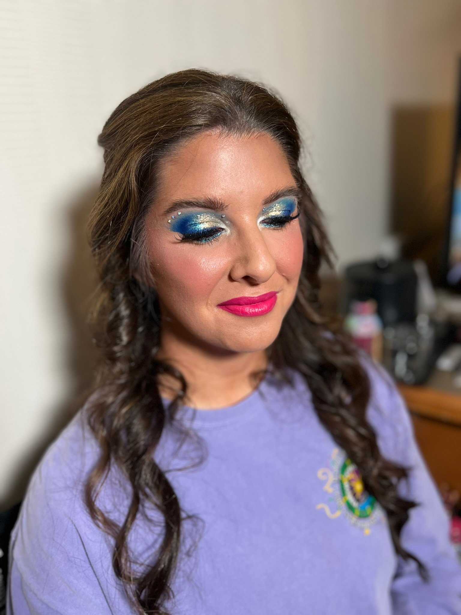Close-up of a woman with styled brown hair and colorful makeup, wearing a purple shirt with a partial rainbow design.