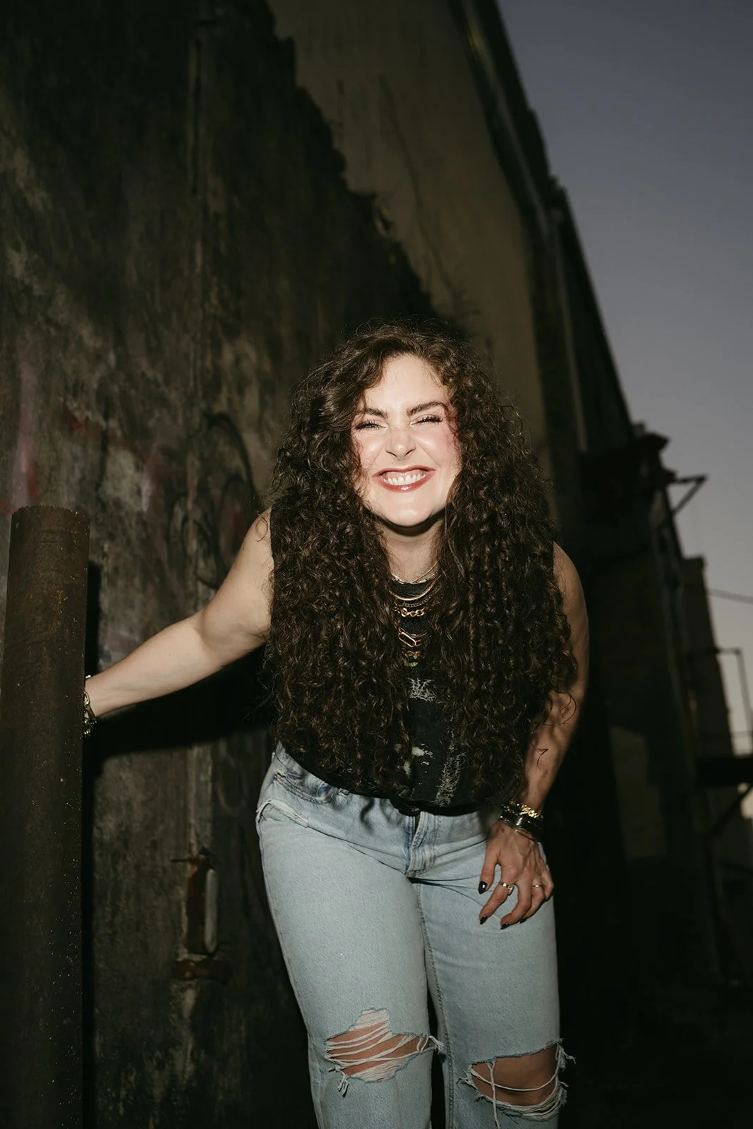 A young woman with long, curly dark hair, smiling and squinting in an outdoor urban setting with a graffiti-covered wall behind her, dressed in a black sleeveless top and ripped light blue jeans.
