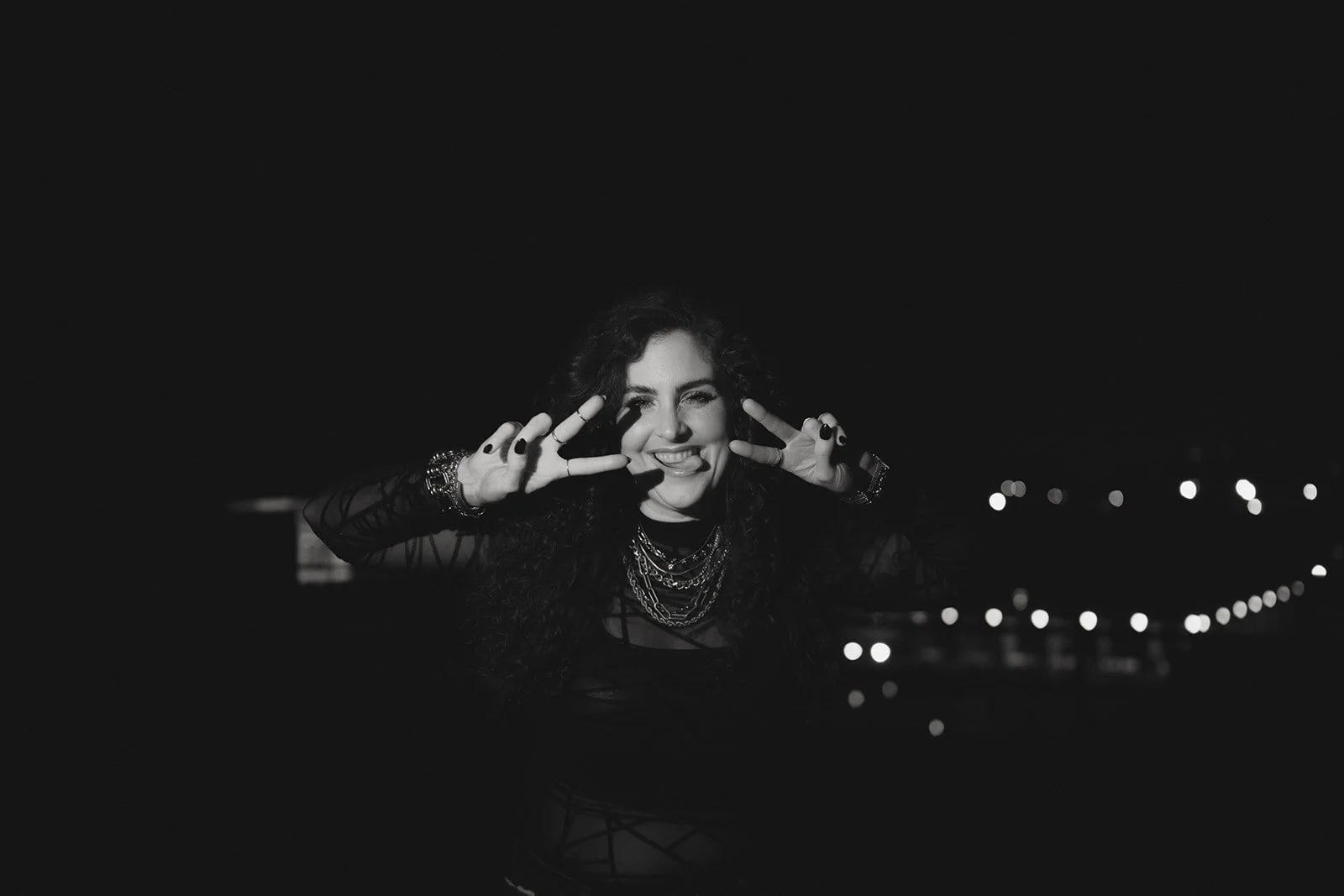 A woman with curly hair making a peace sign with both hands, winking, and sticking out her tongue against a dark background with city lights in the distance.