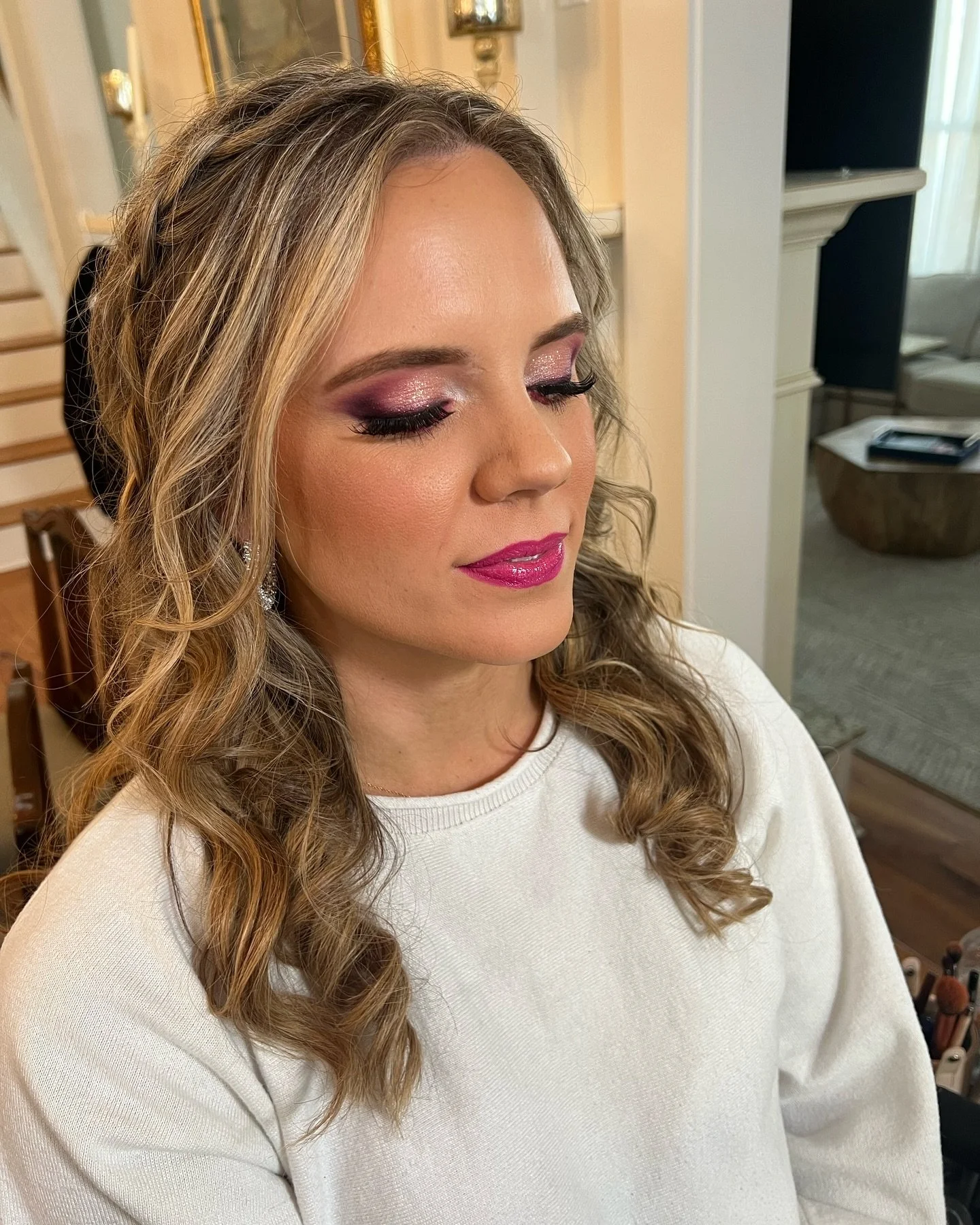 A woman with long, voluminous, curly blonde hair wearing makeup with pink and purple eyeshadow, black eyeliner, long eyelashes, and shiny pink lipstick, is sitting in a room with light-colored walls and furnishings.