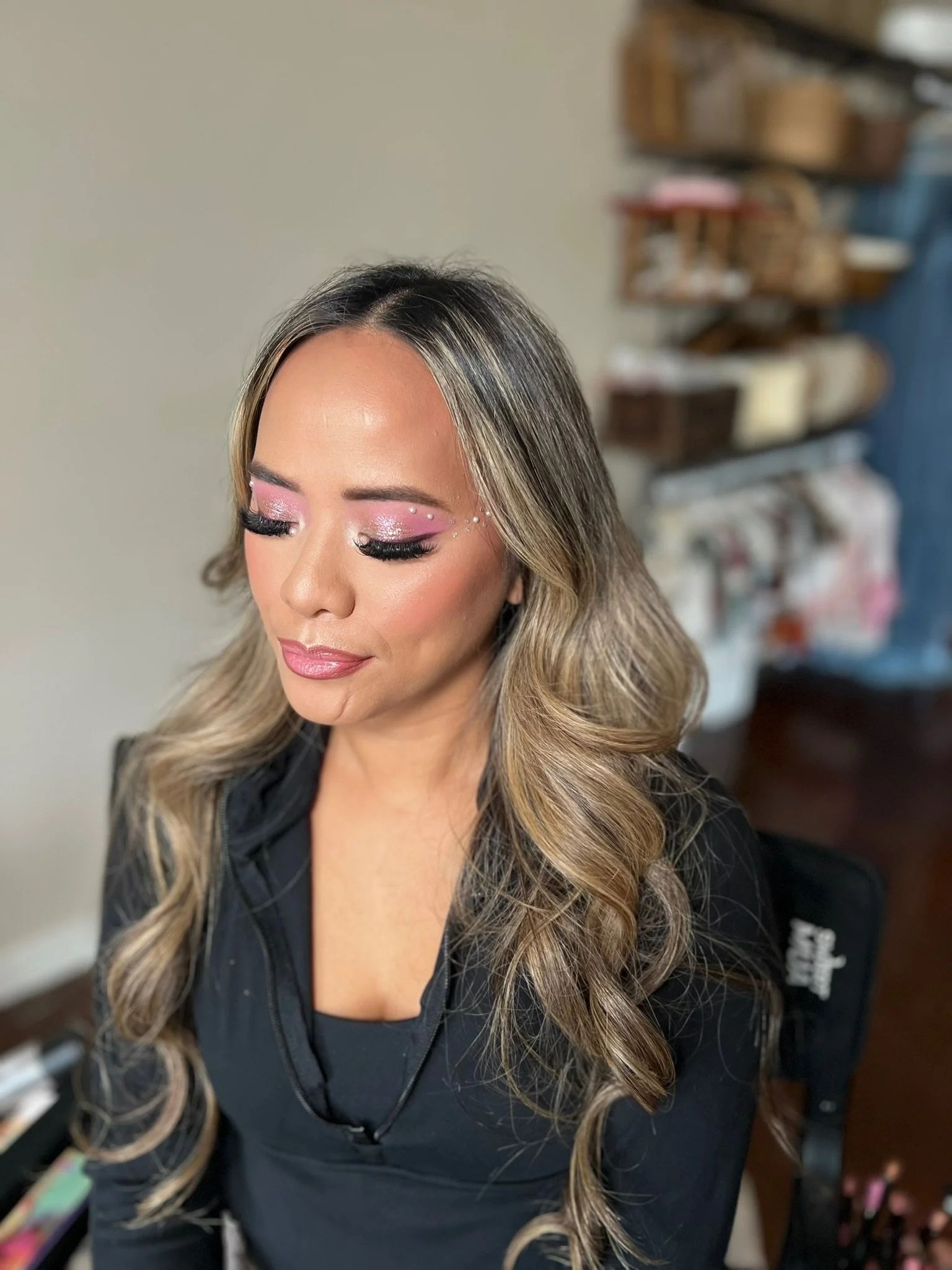 Close-up of a woman with long wavy blonde hair and makeup with pink eyeshadow and rhinestones, with her eyes closed, in a salon or similar setting.