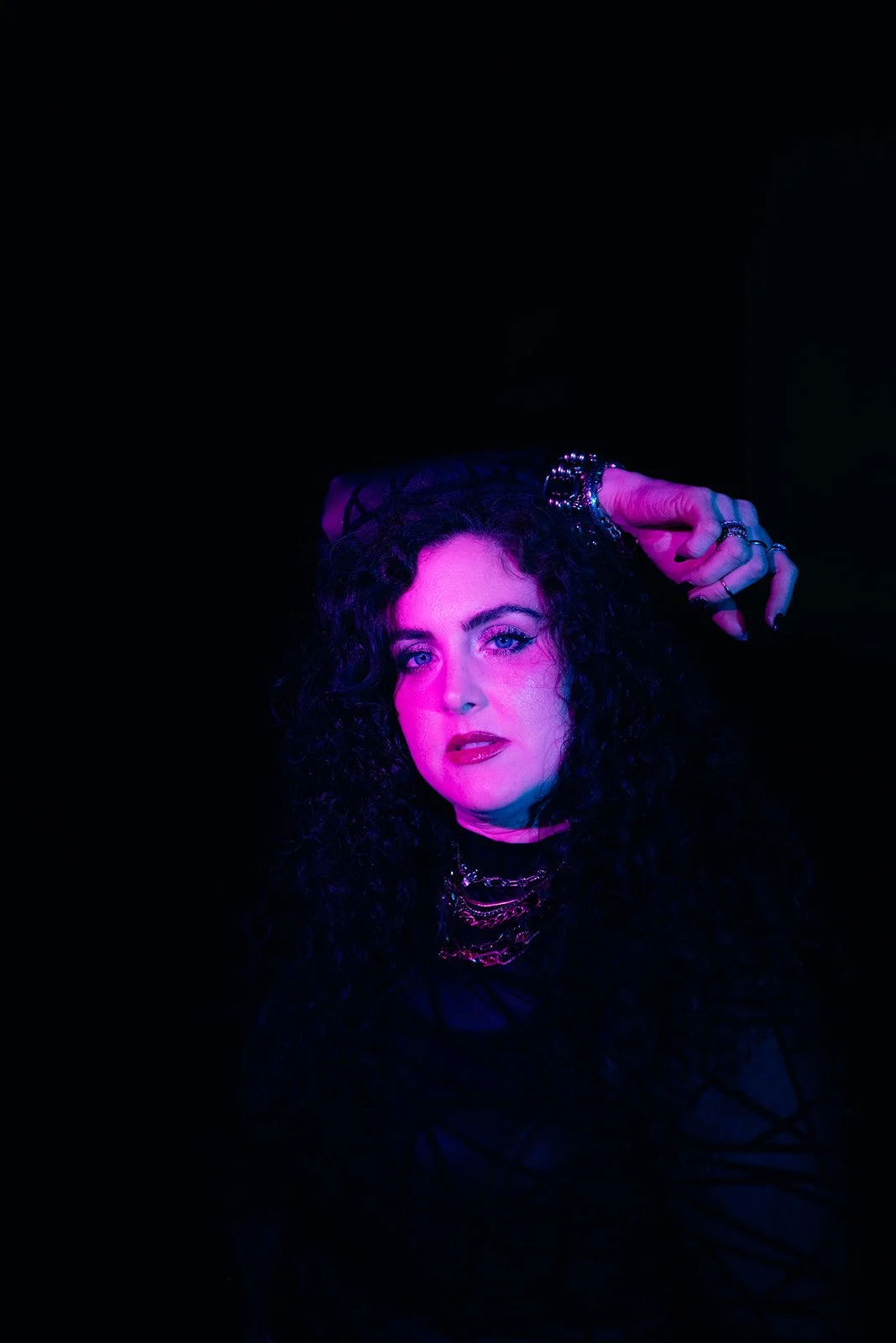 A woman with long, curly black hair and light skin posing in front of a dark background illuminated with purple and pink lighting, wearing jewelry and black clothing.