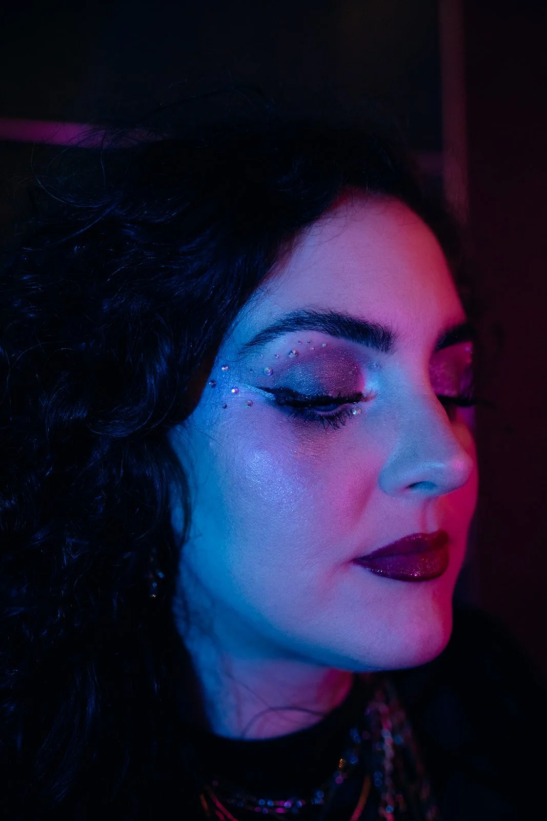 A woman with dark curly hair features vibrant, glittery makeup with rhinestones on her eyelids and bold lipstick, illuminated by colorful lights.