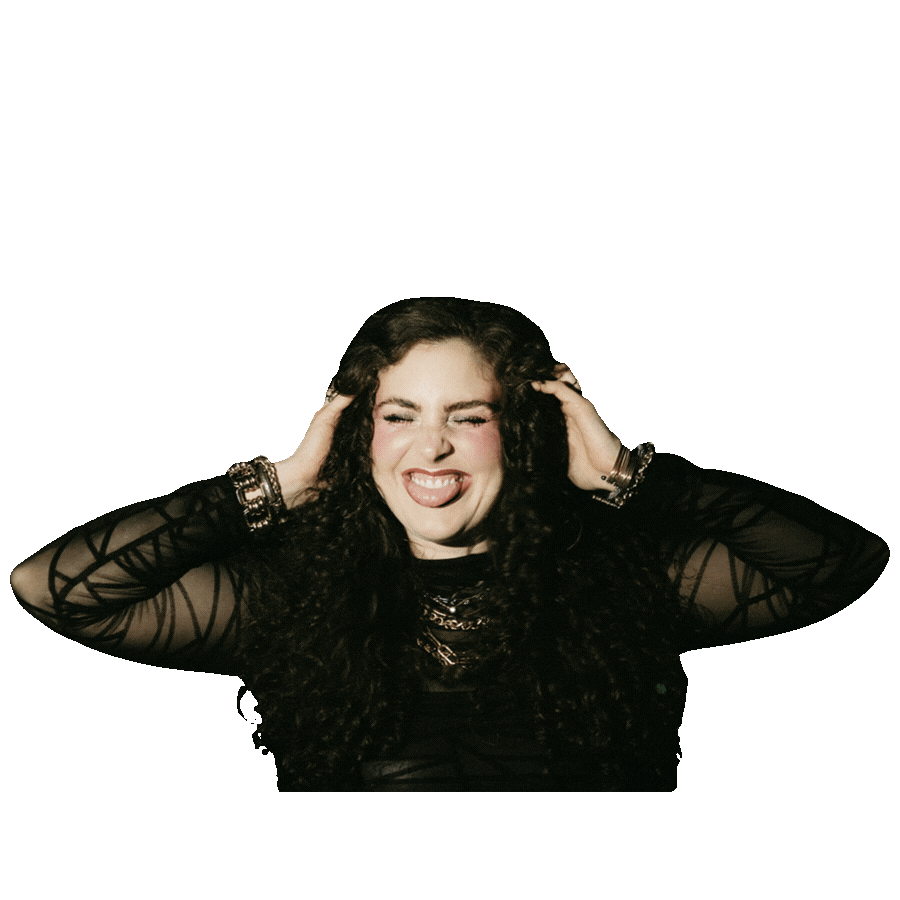 Young woman with curly dark hair, wearing a black sheer top with geometric patterns, making a playful face with tongue out and eyes closed, holding her head with both hands, against a plain white background.