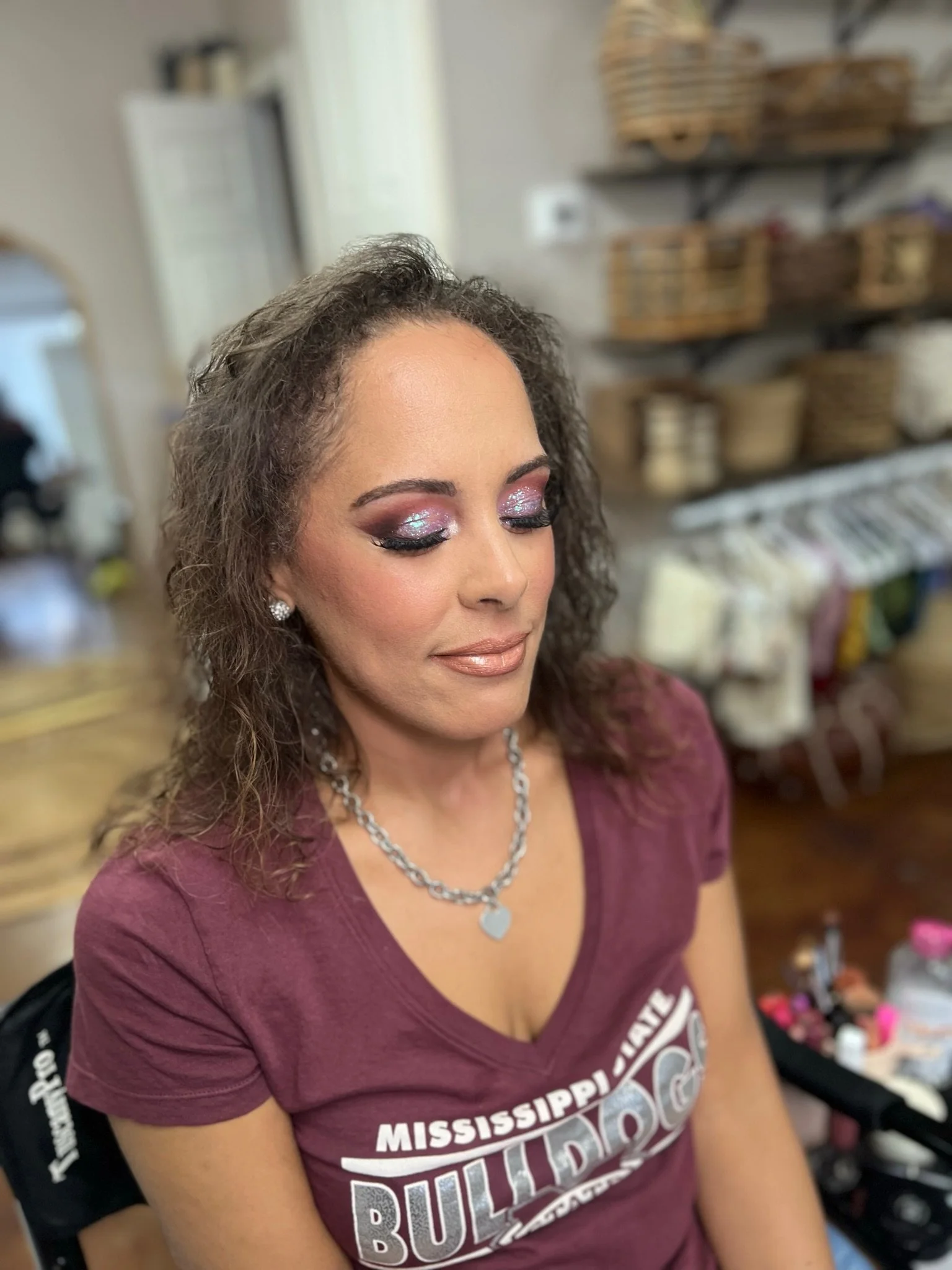 A woman with curly hair wearing makeup and a maroon T-shirt with Mississippi State Bulldogs logo, sitting in a room with shelves of baskets in the background.
