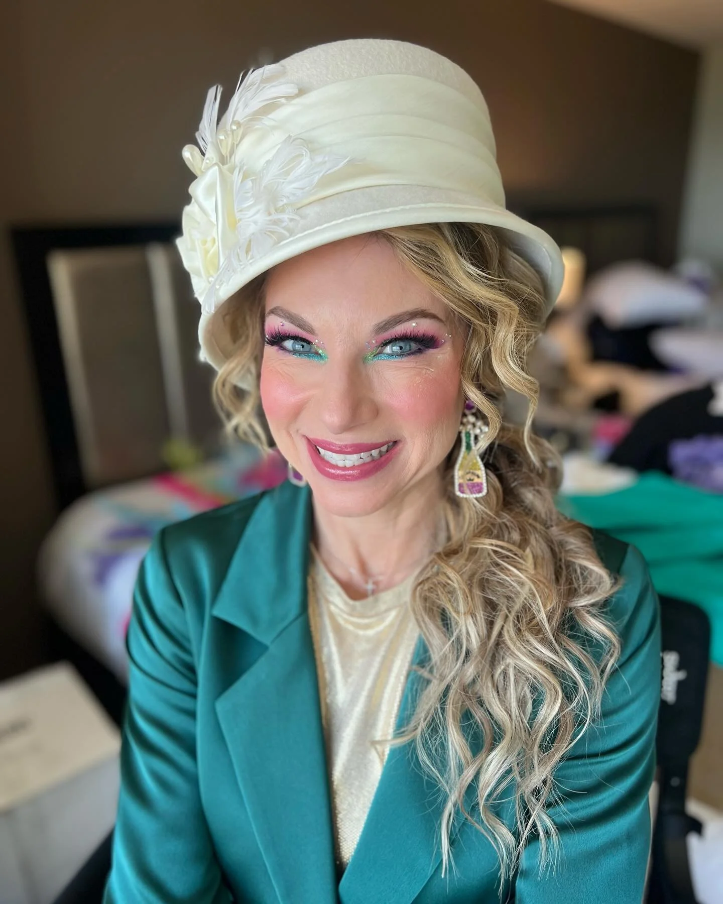 A woman wearing a white hat with feather details, colorful makeup, and a teal blazer, smiling indoors.