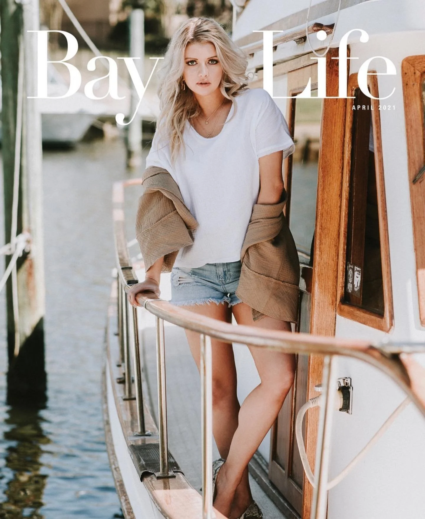 A woman with blonde hair wearing a white t-shirt, denim shorts, and an unbuttoned brown jacket standing on a boat's deck near a harbor with water and boats in the background, looking at the camera.