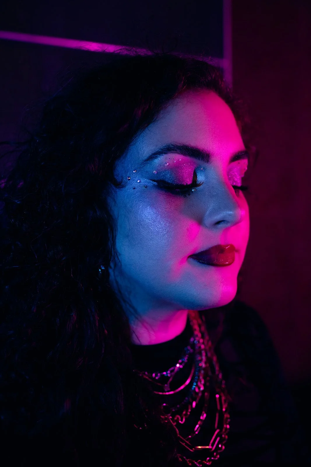 A woman with neon pink and blue makeup, closed eyes, and black lipstick, wearing layered gold chains, illuminated by pink and blue neon lights.