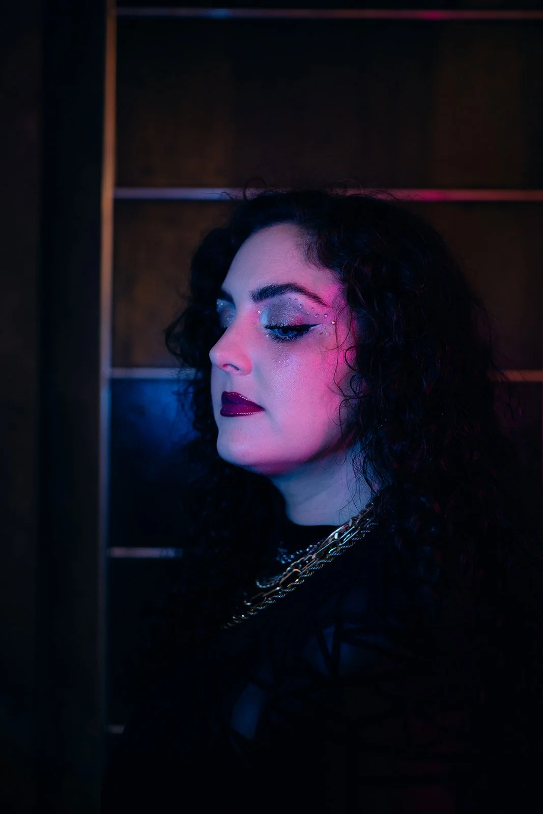 A woman with curly black hair and dramatic makeup, including glittery eyeshadow and dark lipstick, looking to the side in neon-colored lighting.