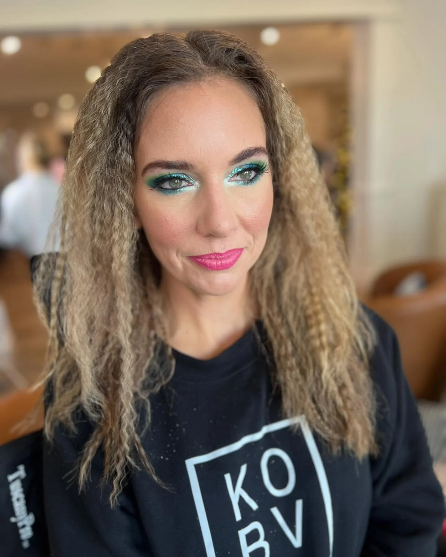 A woman with curly, blonde hair and colorful makeup, wearing a black T-shirt with white text and graphics, indoors with blurred background.