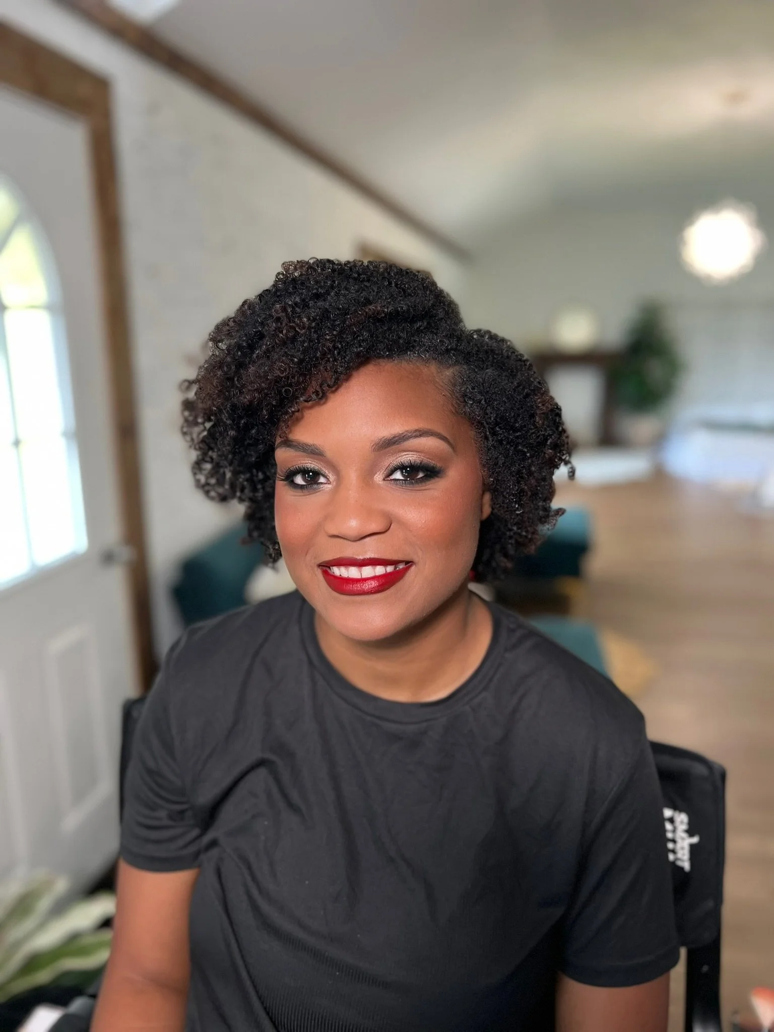 Close-up of a woman with dark, curly hair and red lipstick, smiling, in a living room with a window, green plants, and a wooden shelf in the background.