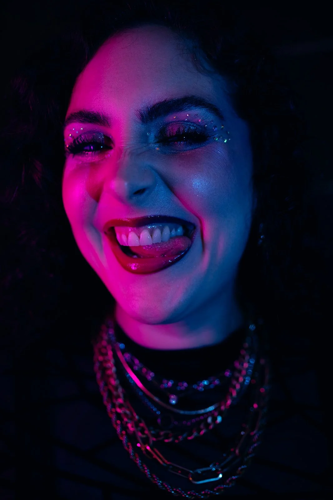 A young woman with dark, curly hair, vibrant makeup with rhinestones around her eyes, sticking her tongue out, wearing multiple necklaces, illuminated by pink and blue neon lighting.