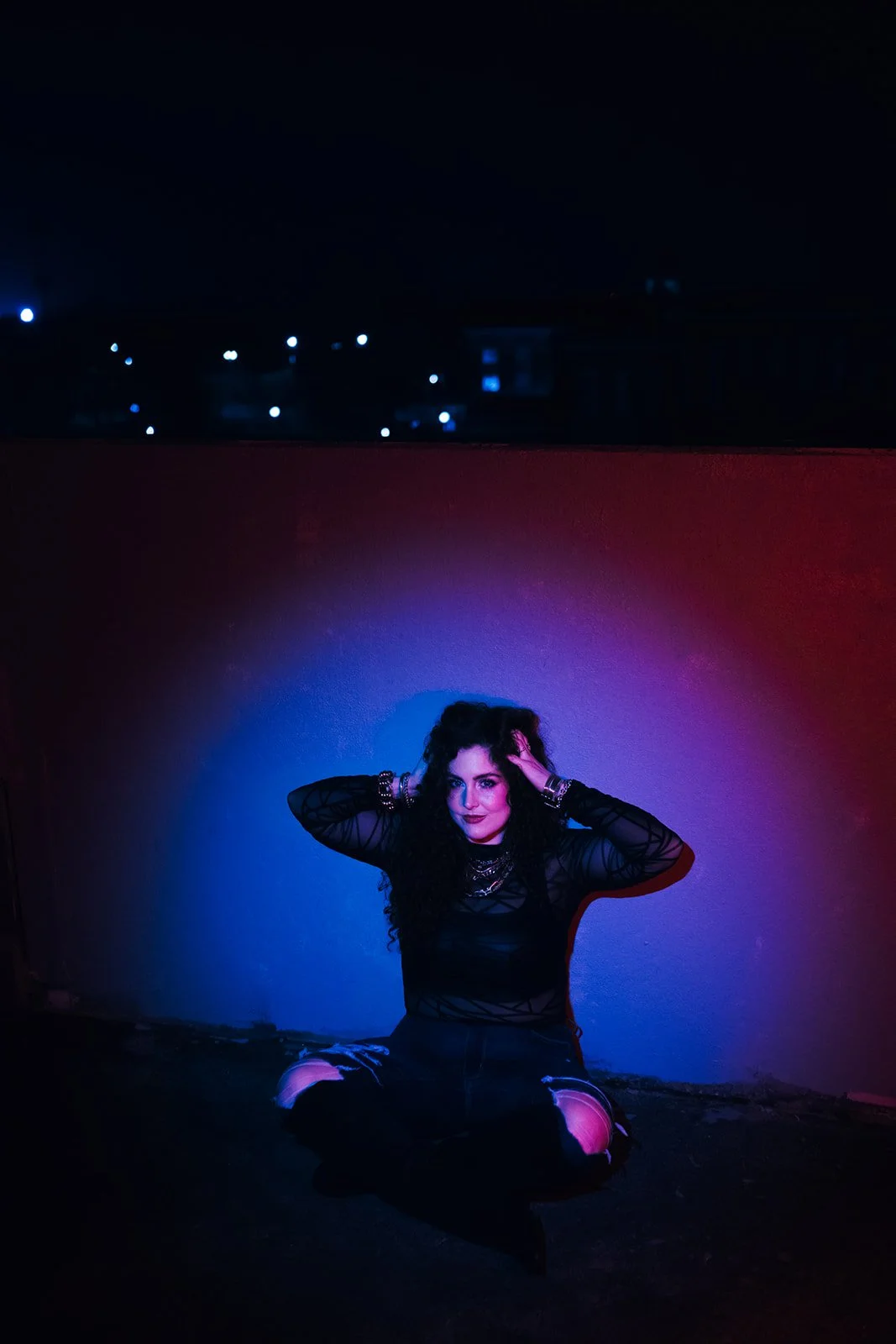 A woman with dark, curly hair sitting on the ground against a wall illuminated by colorful lights, wearing a black sheer top and ripped jeans, with a dark sky and city lights in the background.