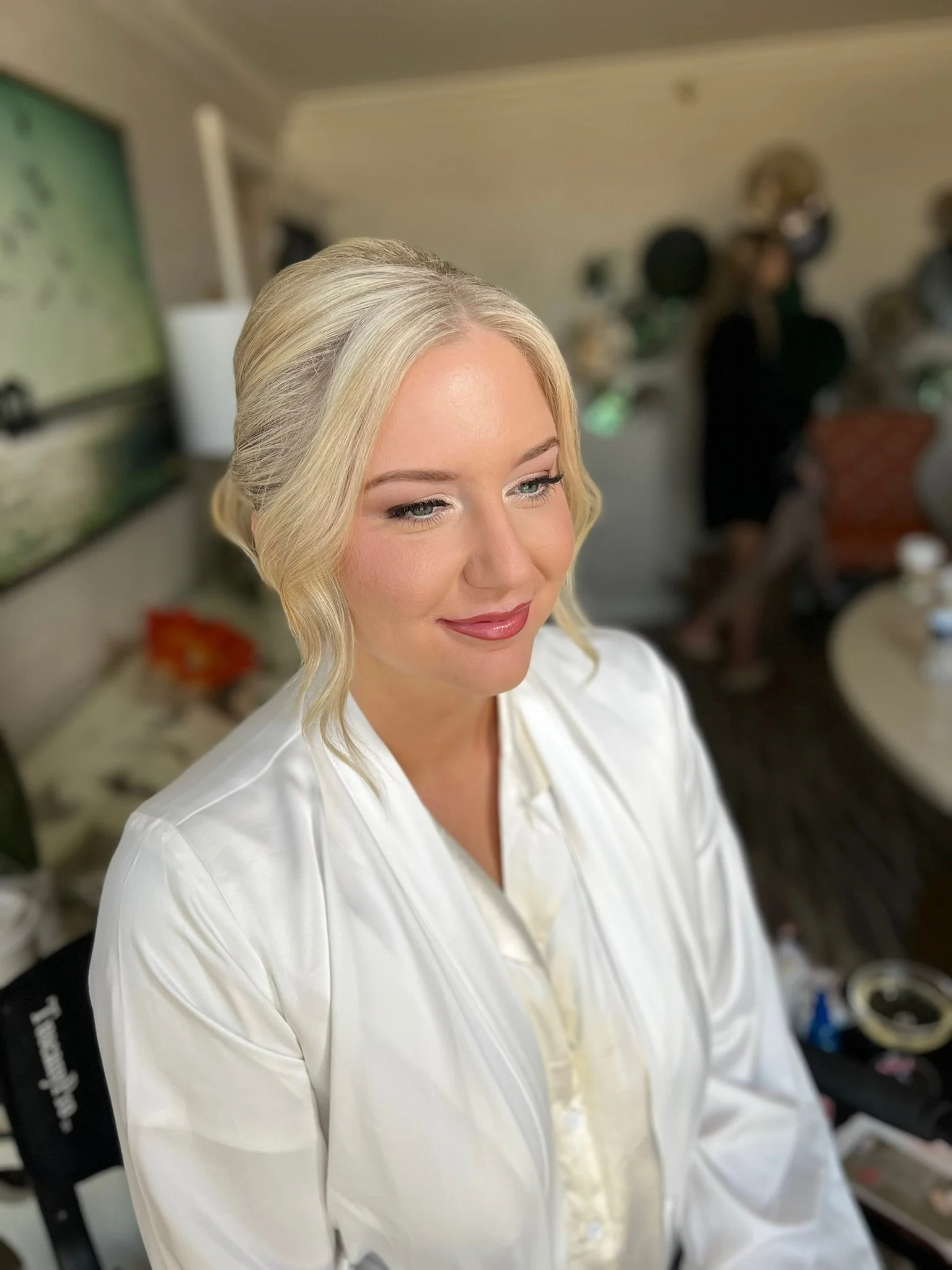 A woman with blonde hair styled in loose waves and wearing a white robe, smiling gently, with a blurred background of people and household items.