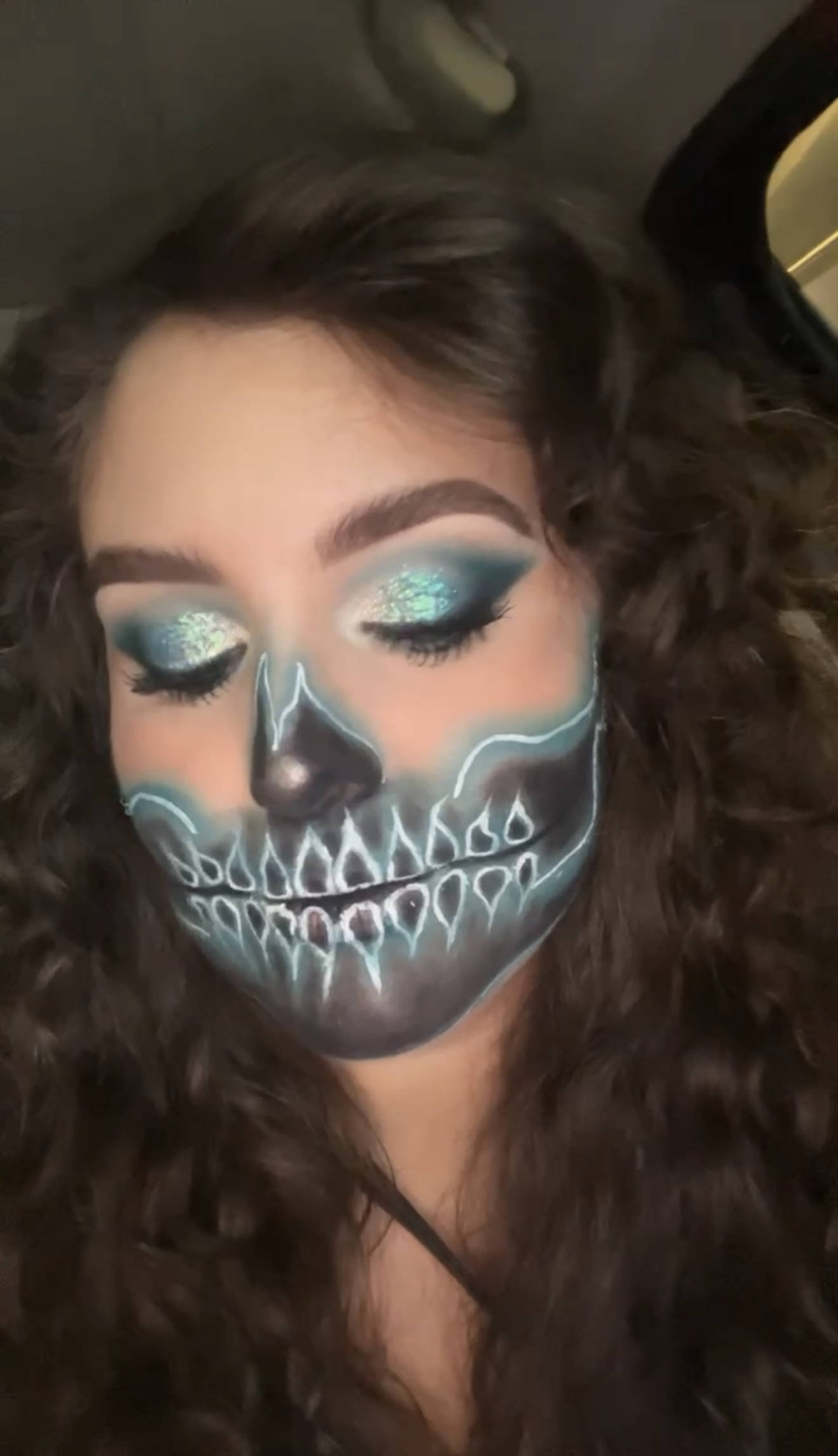 A woman with closed eyes and dark curly hair has detailed face makeup that resembles a colorful skull, featuring turquoise, white, black, and orange hues.