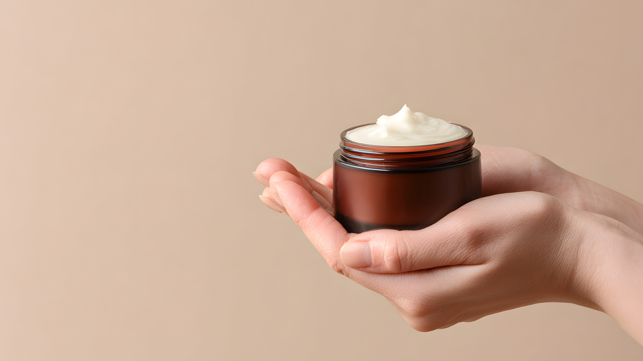 A hand holding an open jar of white cream or moisturizer against a neutral background.