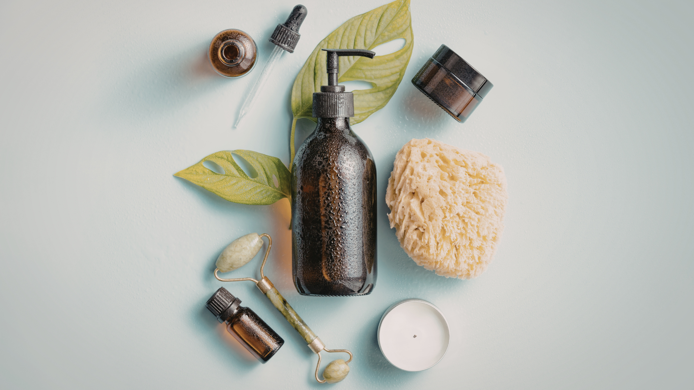 A collection of skincare and beauty products on a light blue surface, including a dark bottle with a pump, a sponge, a small jar, essential oils, and green leaves.