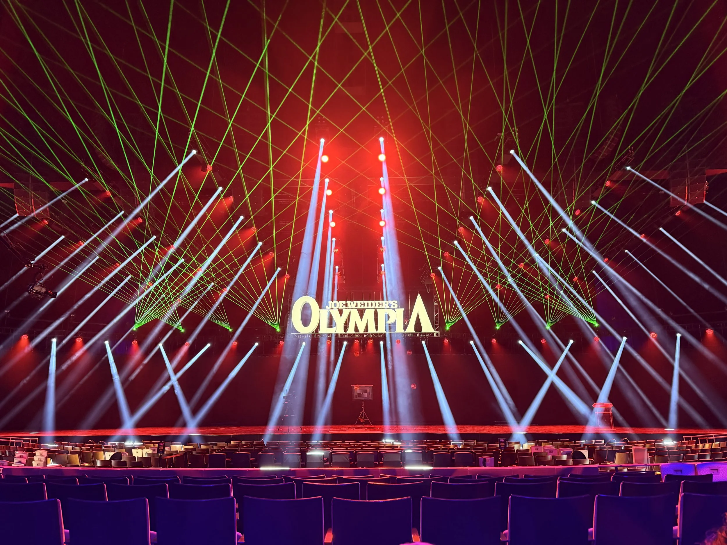 Concert stage with green laser lights, colorful spotlights, and a large sign reading 'Joe Weider's Olympia' at the center. Las Vegas Large Laser Light Show Rental.