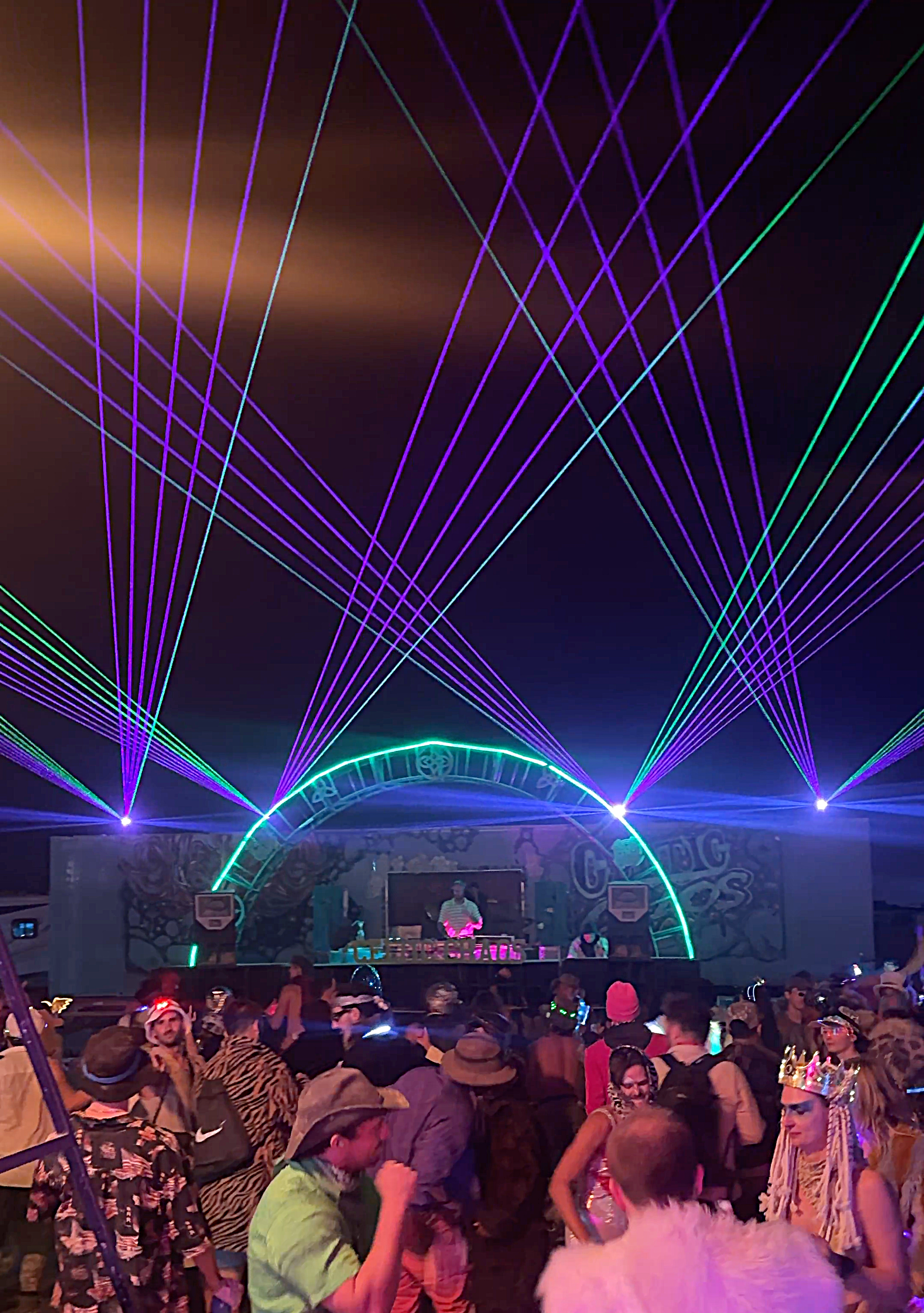 Crowd at an outdoor music festival with a stage featuring a DJ, multiple colorful laser lights, watching a burning man medium laser light show rental.