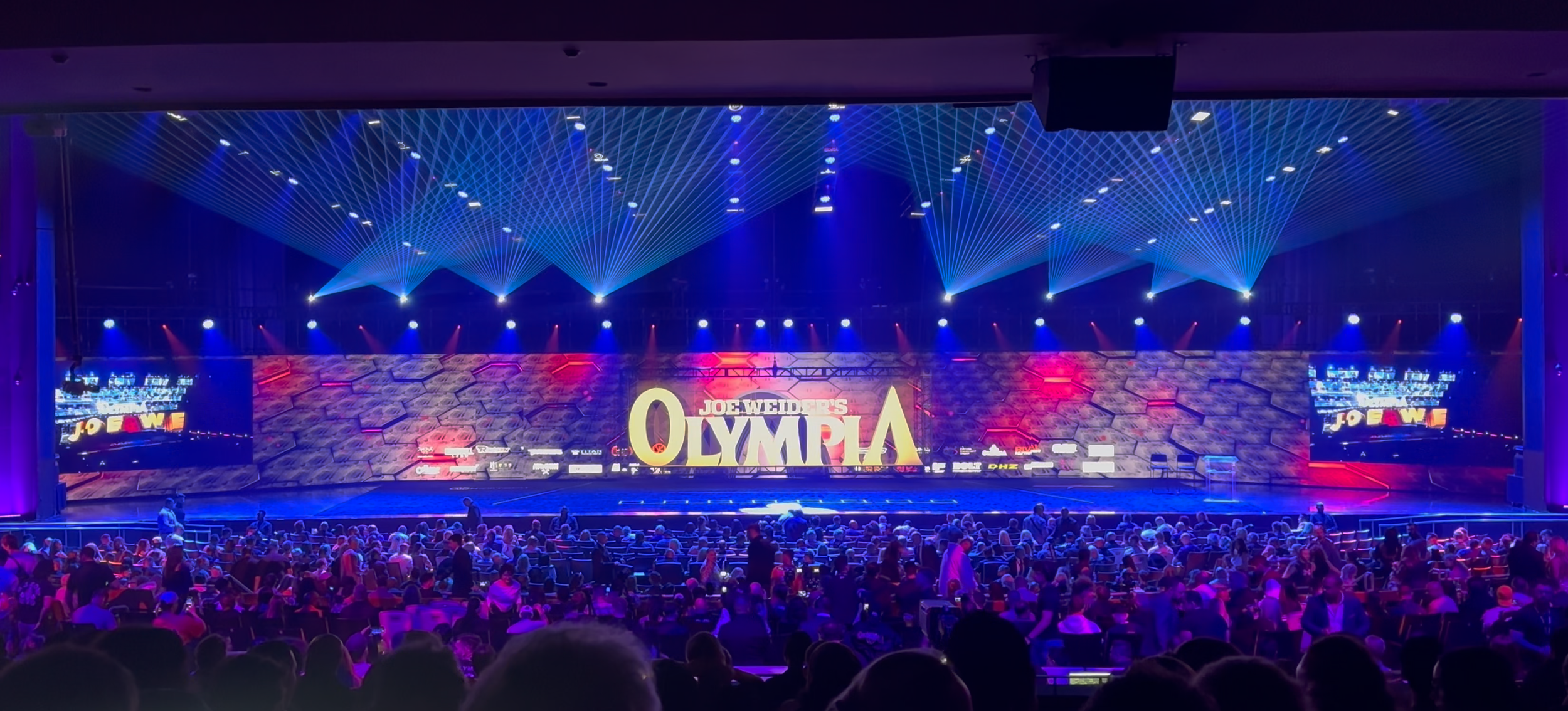 Large indoor stage displaying "Joe Weider's Olympia" and surrounded by an audience. The stage features bright, colorful laser lights and laser light special effects overhead. Las Vegas corporate laser light show rental.