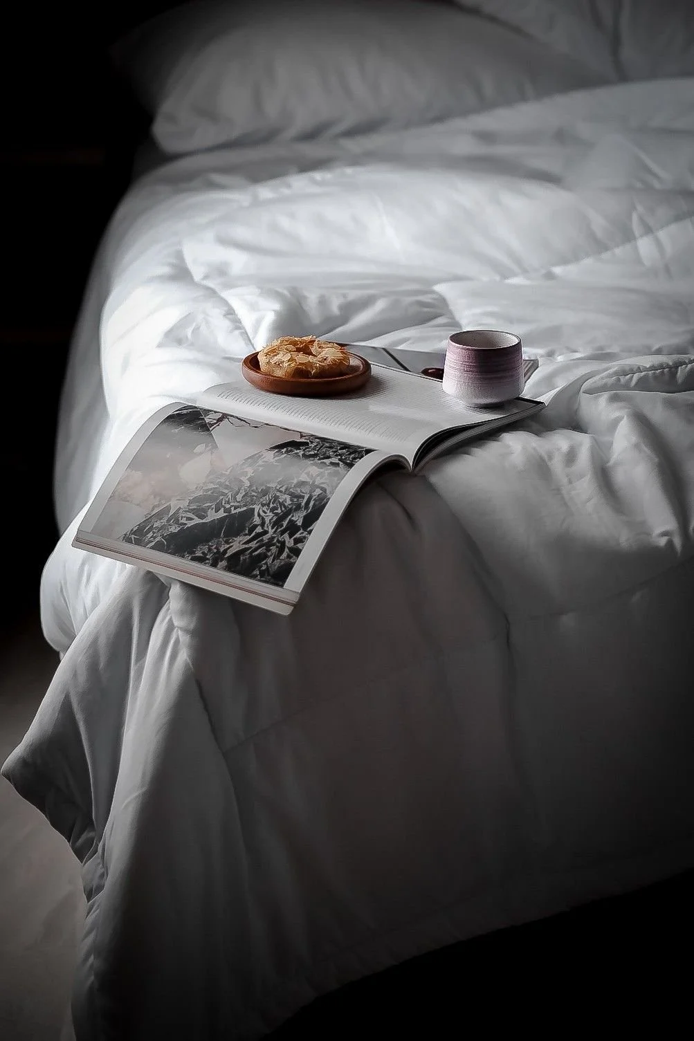 A neatly made bed with white bedding, a cup, an open book, a magazine, and a plate of cereal on top, with soft lighting casting shadows.