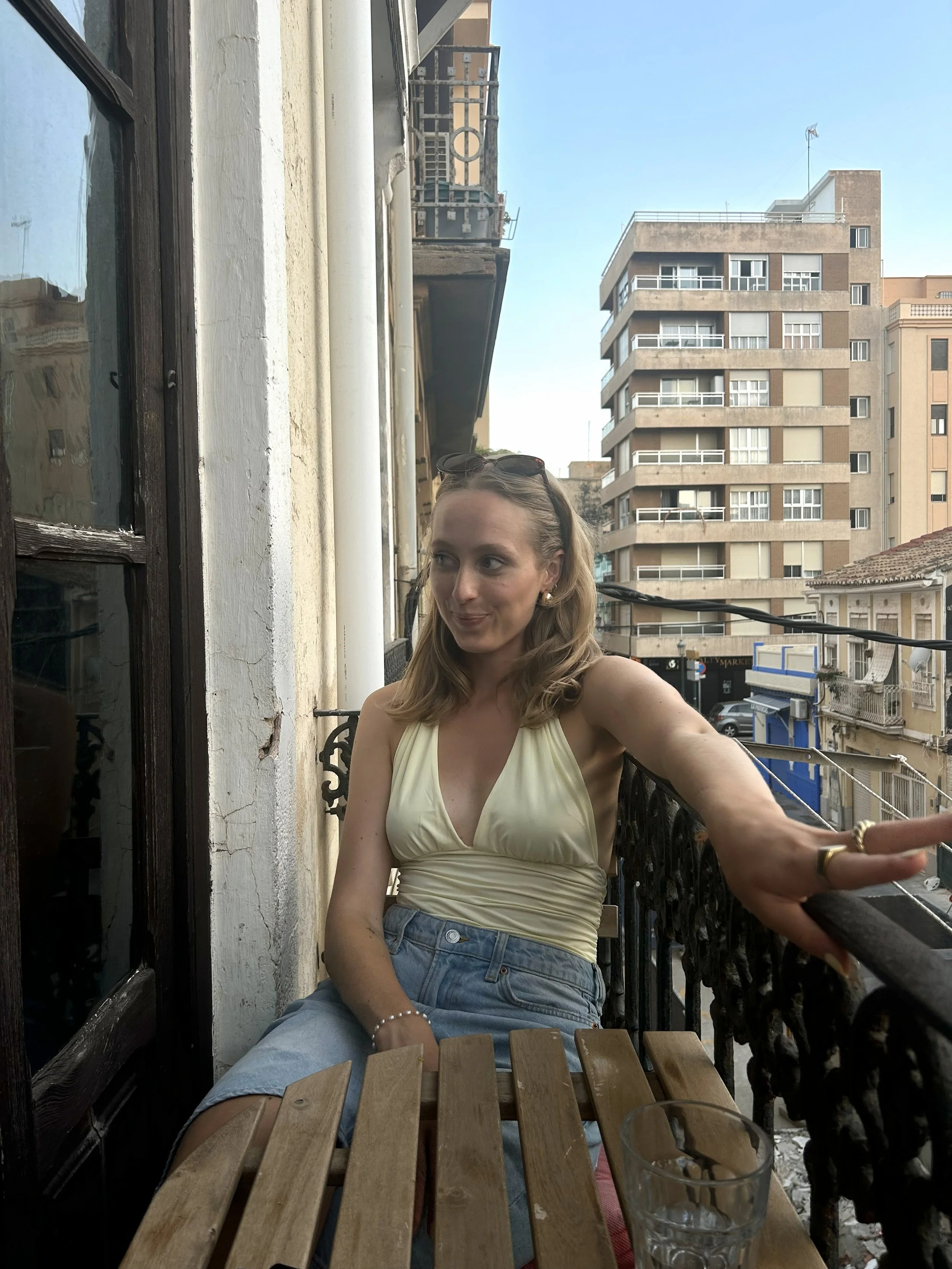 A young woman sitting on a balcony with a cityscape in the background. She is wearing a yellow sleeveless top, denim shorts, and sunglasses on her head. She is reaching out with her right arm, possibly for a selfie or to reach for something, with a slight smile on her face. There is a glass on the table in front of her.
