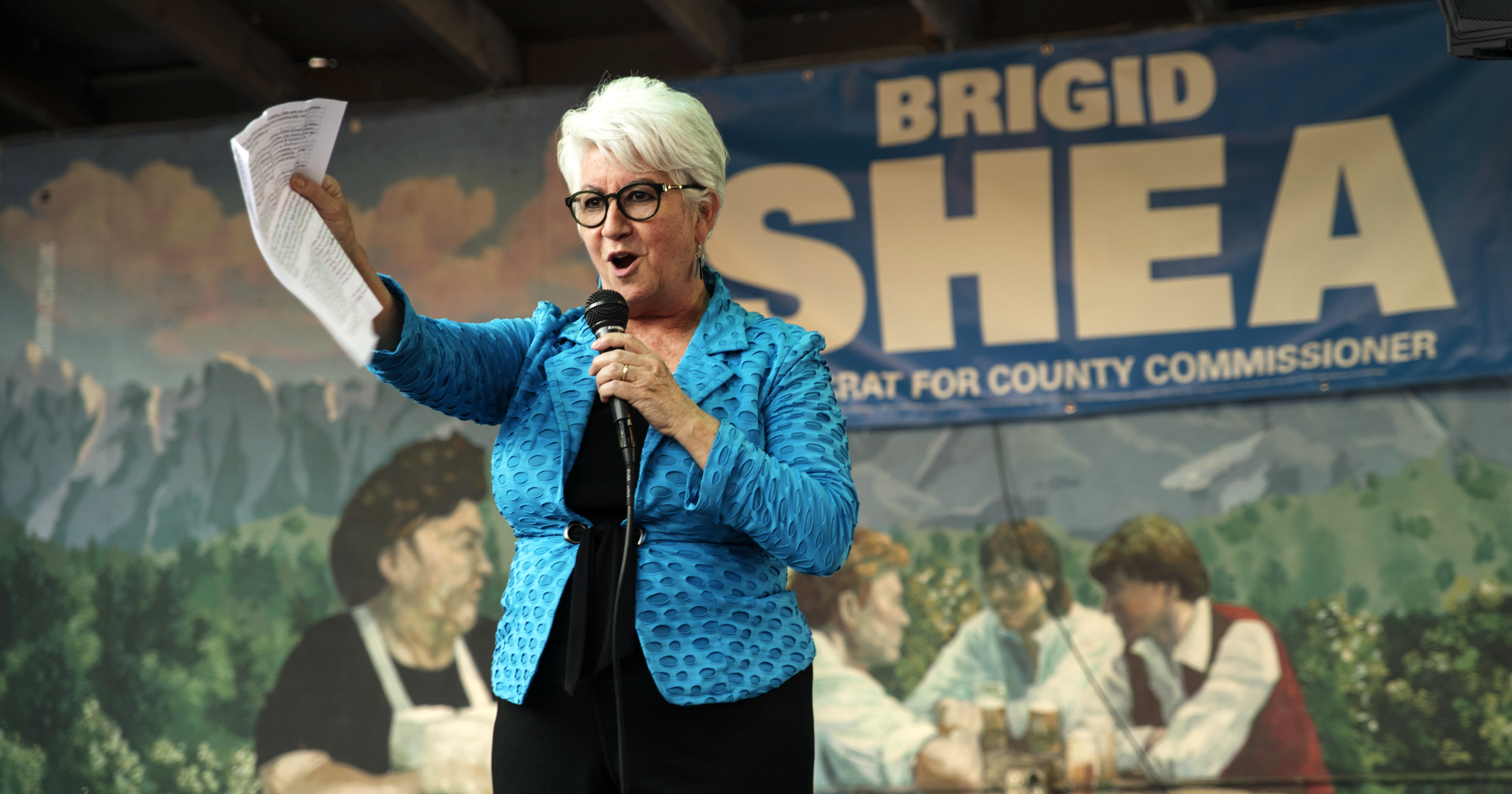 A woman with white hair, glasses, and a blue textured jacket is speaking into a microphone and gesturing with her left hand. She is standing in front of a banner with the words 'BRIGID SHEA' and 'DEMOCRAT FOR COUNTY COMMISSIONER.' The background shows a painted mural of mountains and people sitting at a table outdoors.