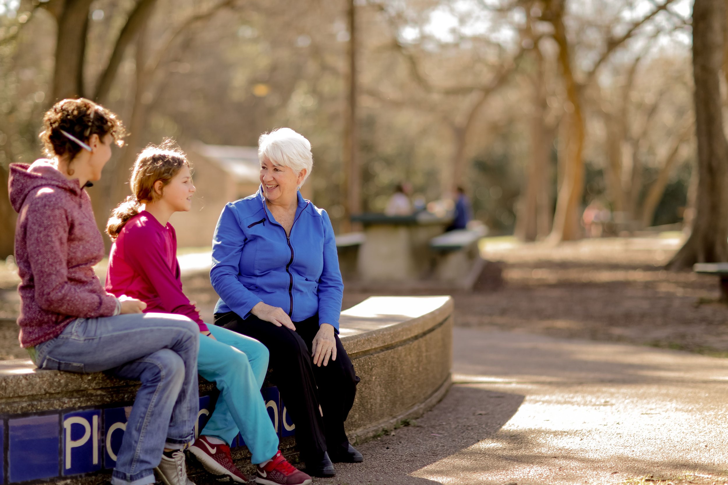 An elderly woman and two young girls sitting on a park bench, engaging in conversation. The scene is outdoors with trees and a sunny, clear day.