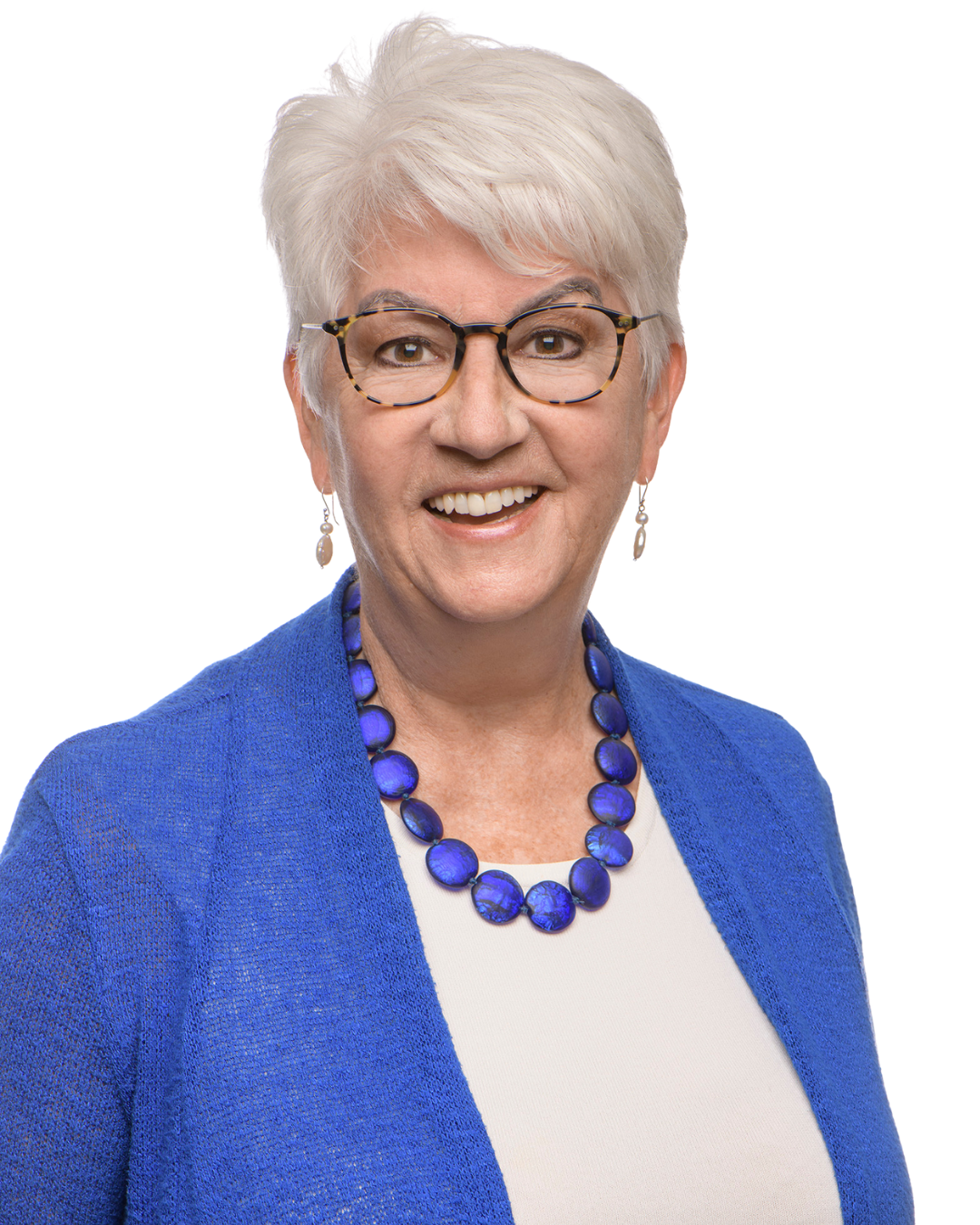 A middle-aged woman with short white hair and glasses, wearing a blue necklace, a white top, and a blue blazer, smiling at the camera.