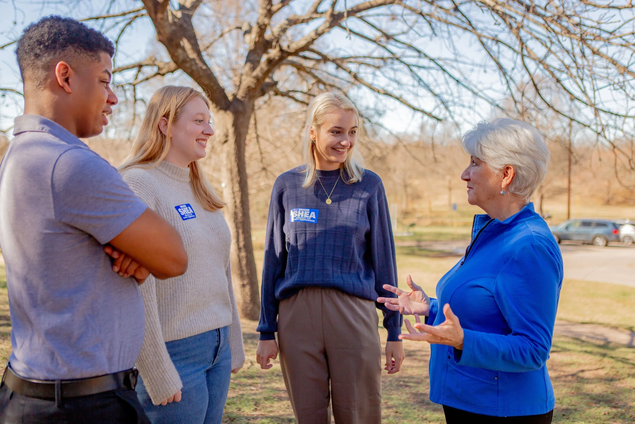 Four women standing outdoors, engaging in conversation, with trees and parked cars in the background, during daytime. One woman is speaking to three others.