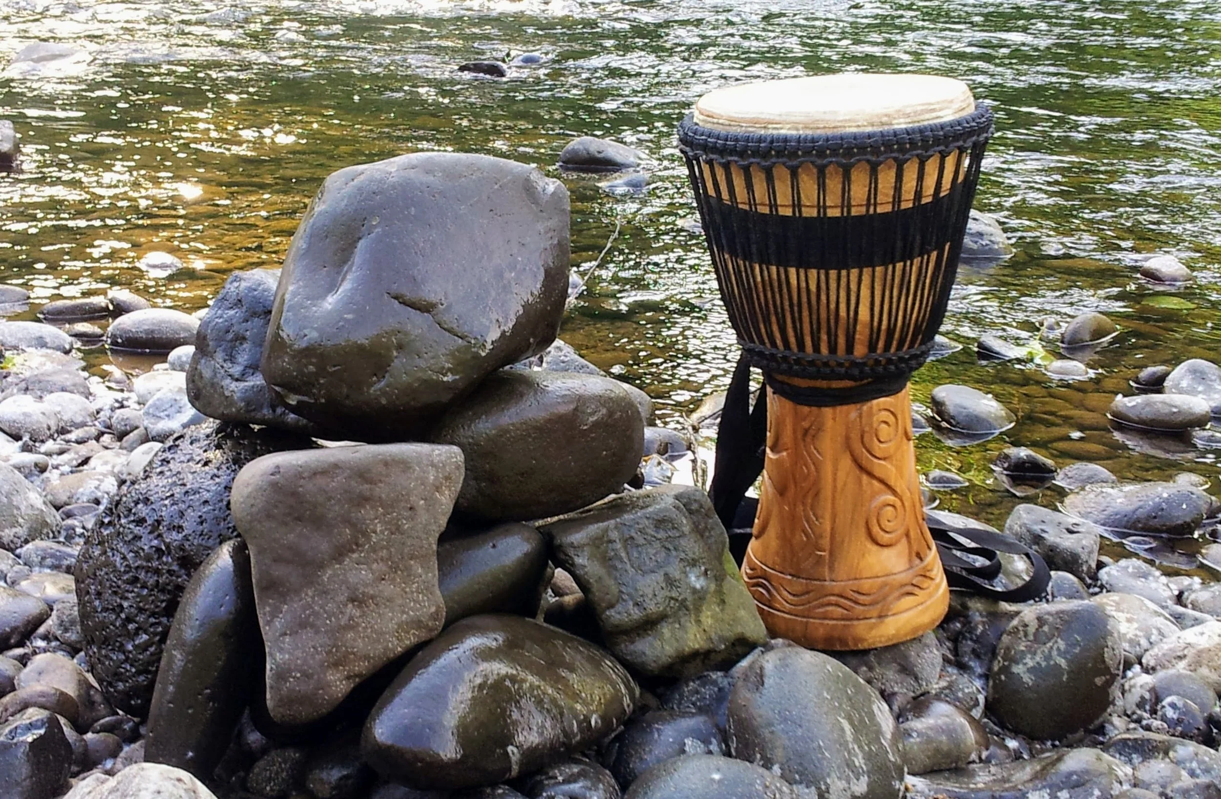 A djembe drum placed on a rocky riverbank near water with small rounded stones and larger rocks alongside.