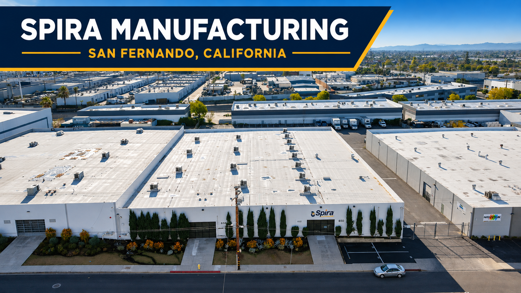 Aerial view of Spira manufacturing building in San Fernando, California, featuring a large white flat roof and surrounding landscape with trees and parked vehicles.