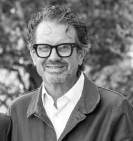 Black and white photo of a smiling man with glasses, curly hair, and a beard, wearing a collared shirt and jacket outdoors with blurred trees in the background. Ian Coats MacColl