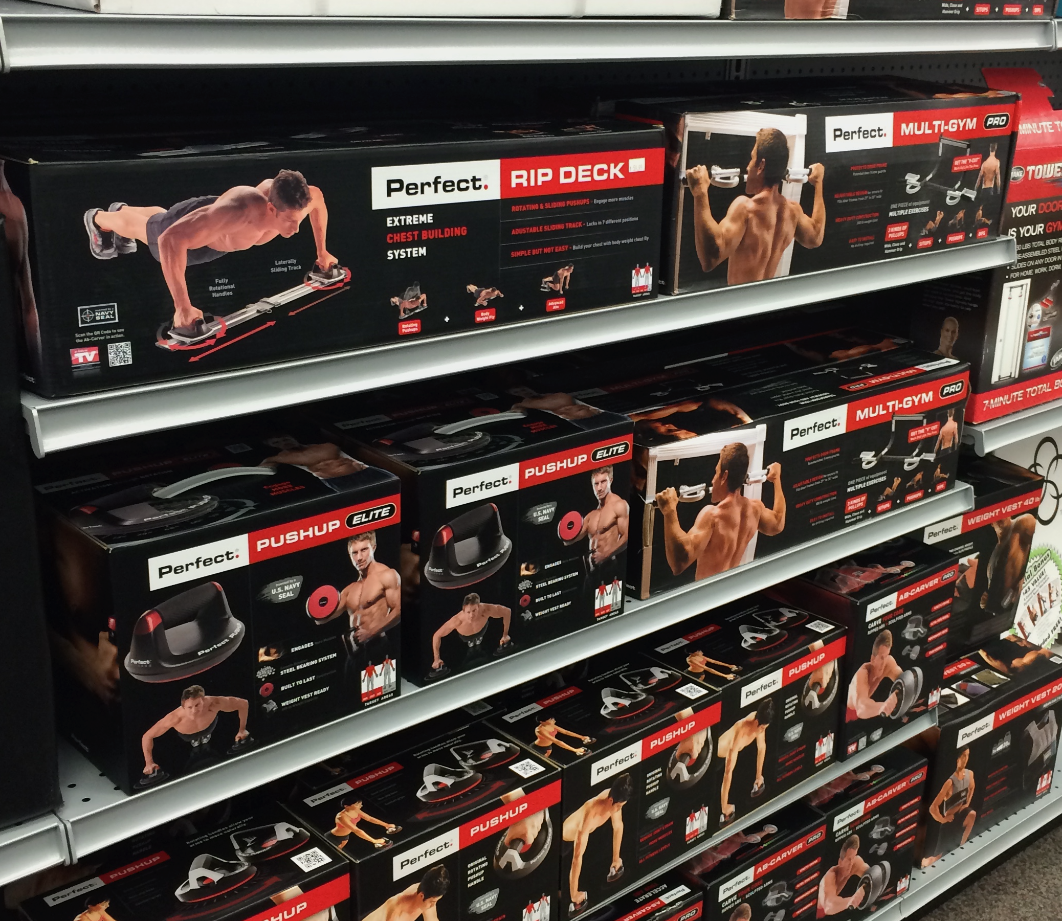 Perfect Fitness boxes in retail. Shelves of exercise equipment including push-up bars and multi-gym systems with images of men demonstrating exercises.